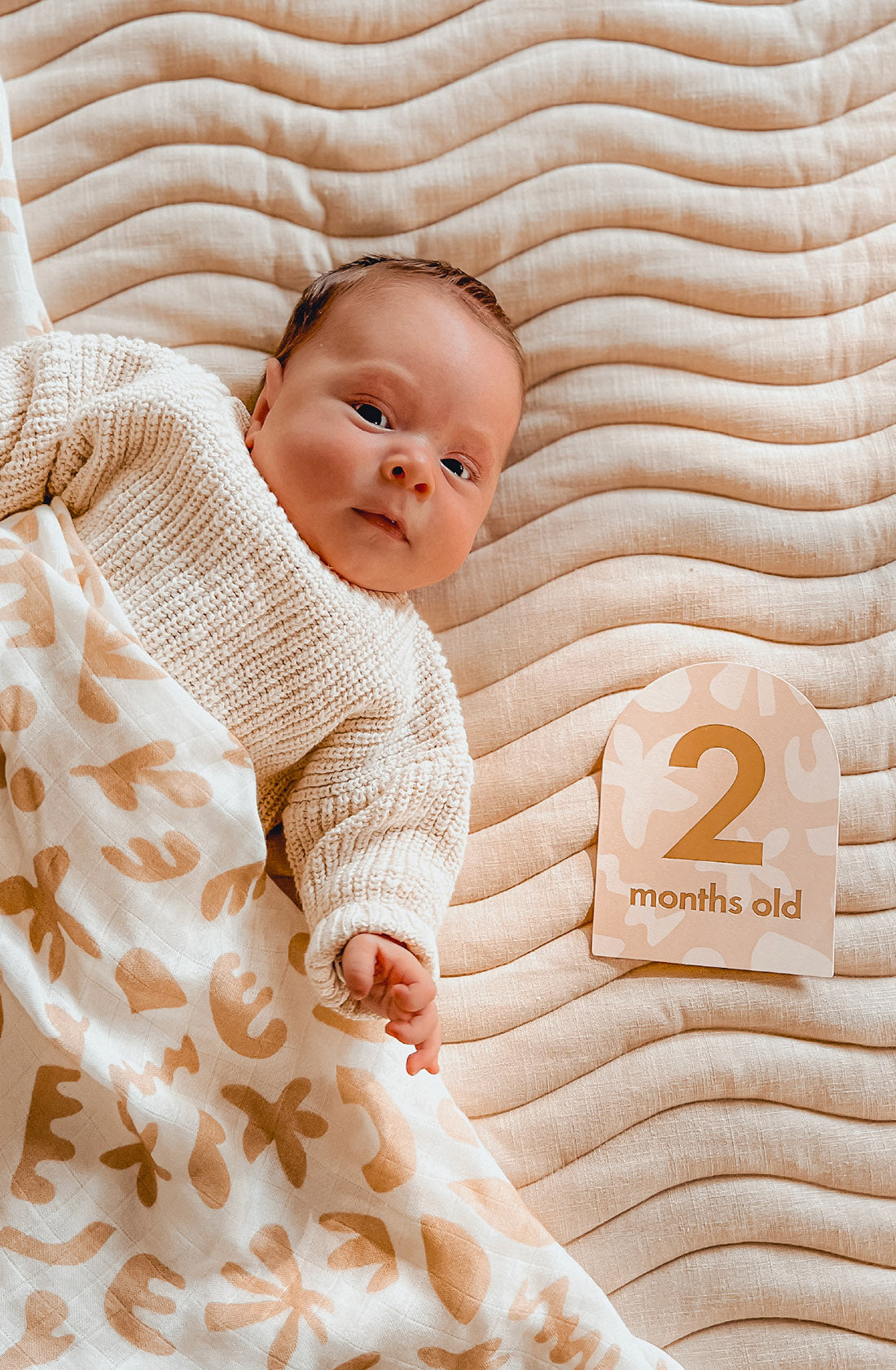 A baby in a cream knit sweater lies on a beige blanket next to a FOX & FALLOW - Baby Milestone Cards Helios card that reads "2 months old.
