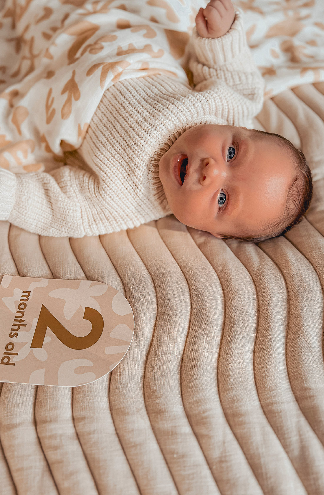 A smiling baby in a cream sweater lies on a beige patterned blanket, partly covered by a knit. Beside them is a FOX & FALLOW - Baby Milestone Cards Helios card that says “2 months old.”.