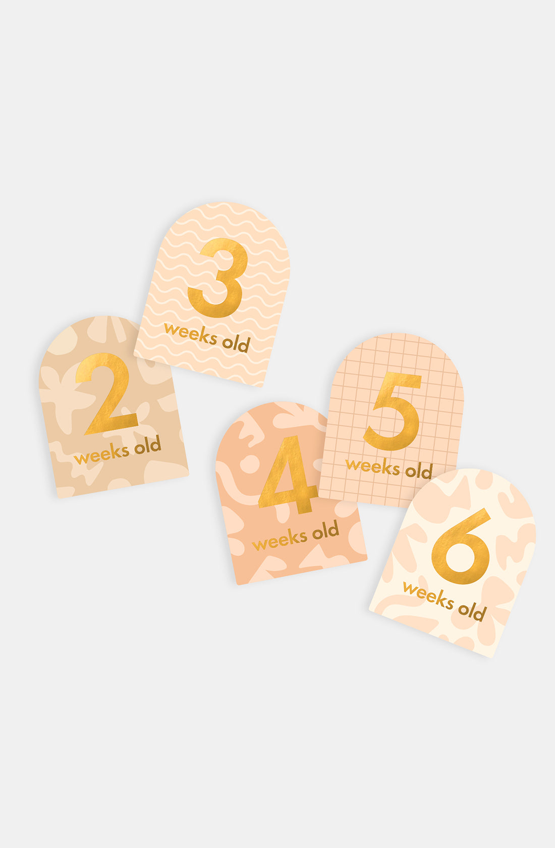 Five pastel-patterned FOX & FALLOW Baby Milestone Cards Helios, with gold numbers and "weeks old" text (numbers 2–6), are fanned out and ideal for birth announcements or tracking your baby's growth.