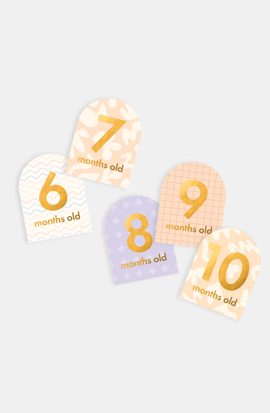Five "6 months old" to "10 months old" milestone cards from the FOX & FALLOW Helios collection showcase pastel patterns and gold lettering, elegantly arranged on a light background.