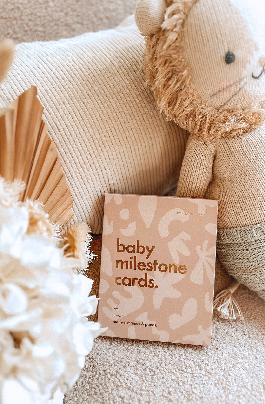 A box of FOX & FALLOW Baby Milestone Cards Helios sits on a beige cushion, next to a plush lion toy and dried flowers in a cozy, neutral-toned space.