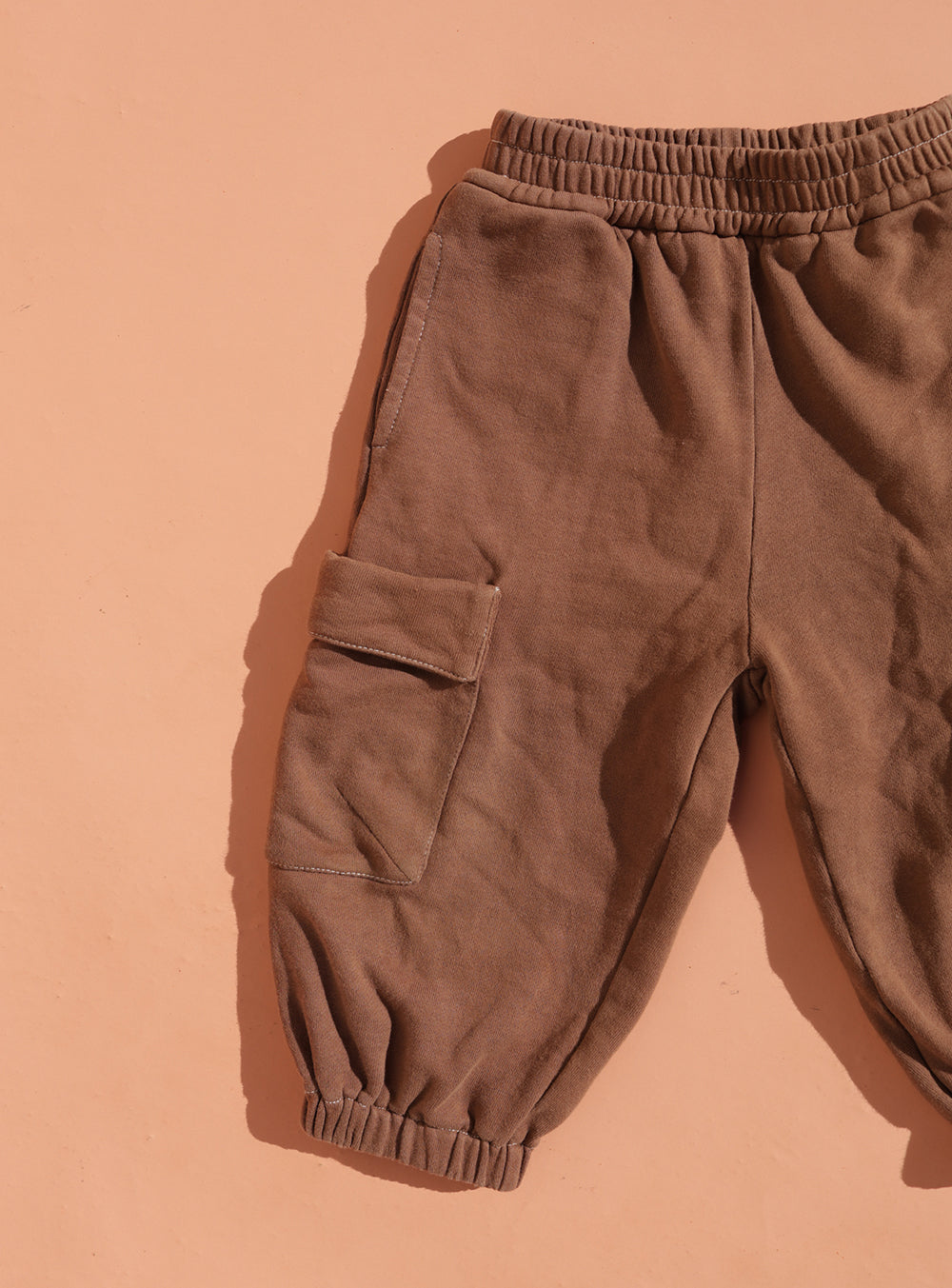 The SMALL SWIM CLUB Small Swim Club - Choccy Cargo Trackies, brown brushed cotton with an elastic waistband and cuffs, are laid flat on a peach surface, showing the left leg's large cargo pocket and oversized fit.
