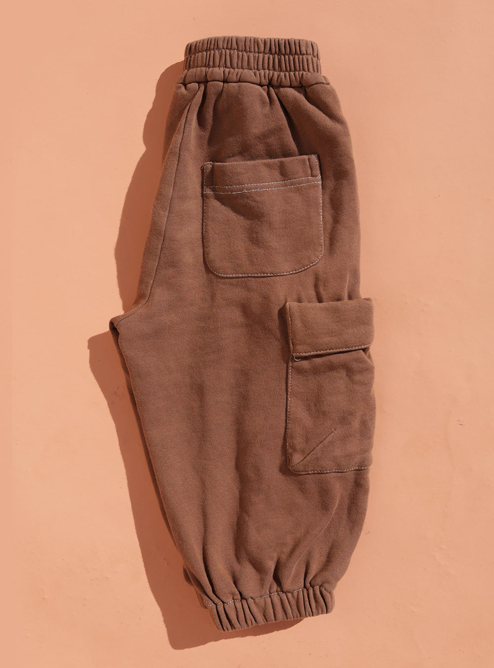 The Small Swim Club - Choccy Cargo Trackies by SMALL SWIM CLUB, made from soft brushed cotton with an elastic waistband, cuffs, front patch pocket, and side cargo pockets, are shown laid flat on a peach-colored surface.