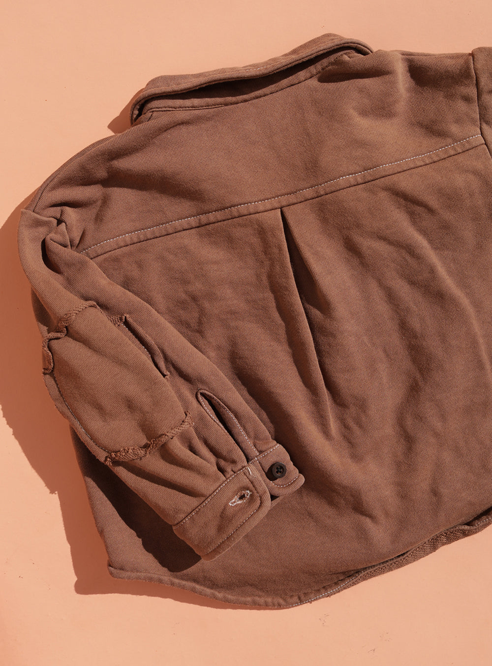 The SMALL SWIM CLUB Choccy Cargo Jacket in brown brushed cotton features a pleated back and visible stitching on the elbow patch, shown flat on a peach background. Pair it with matching pants for a true utility look.