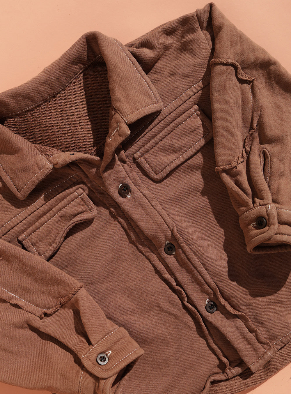 The SMALL SWIM CLUB Small Swim Club - Choccy Cargo Jacket, a brown brushed cotton button-up with front pockets and rolled sleeves, is displayed flat on a peach surface, capturing a relaxed utility style.