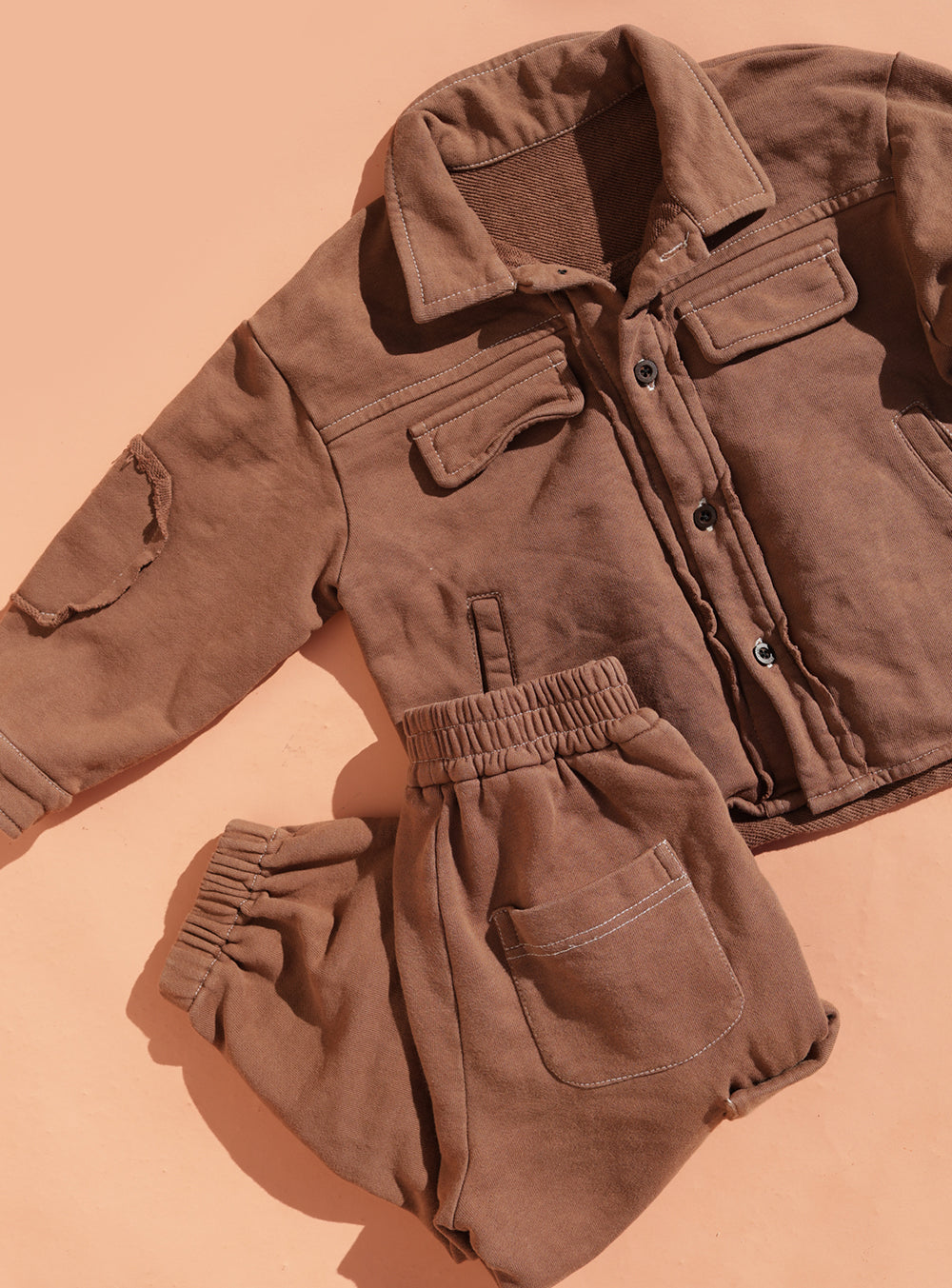 The Small Swim Club - Choccy Cargo Jacket by SMALL SWIM CLUB is shown laid flat on a peach background, featuring visible stitching, patch pockets, and button details for a stylish utility look.