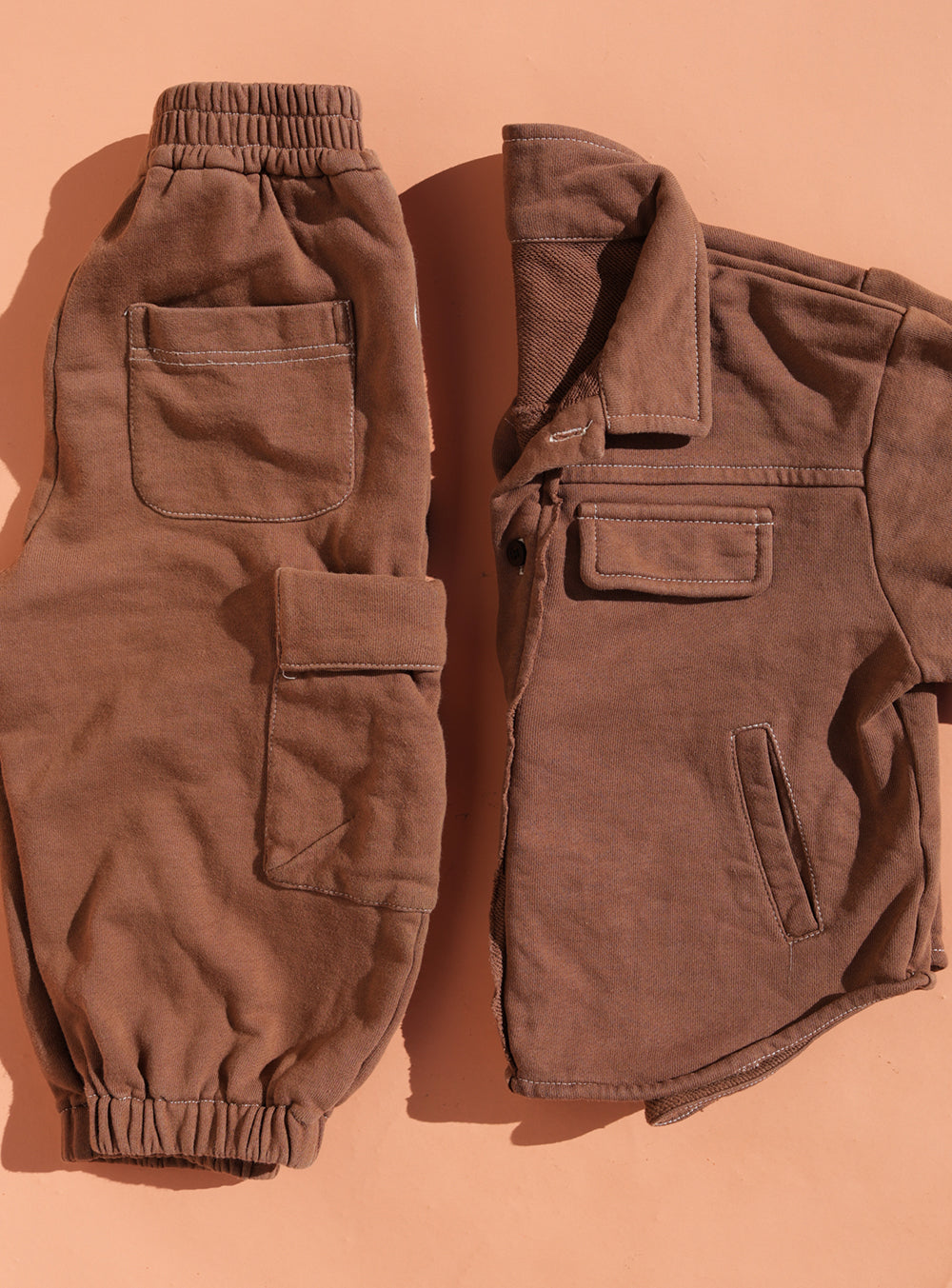 The SMALL SWIM CLUB Choccy Cargo Jacket is a brown brushed cotton button-up with utility pockets, shown laid flat on a peach-colored surface. Matching cargo pants with elastic cuffs and multiple pockets complete the casual set.