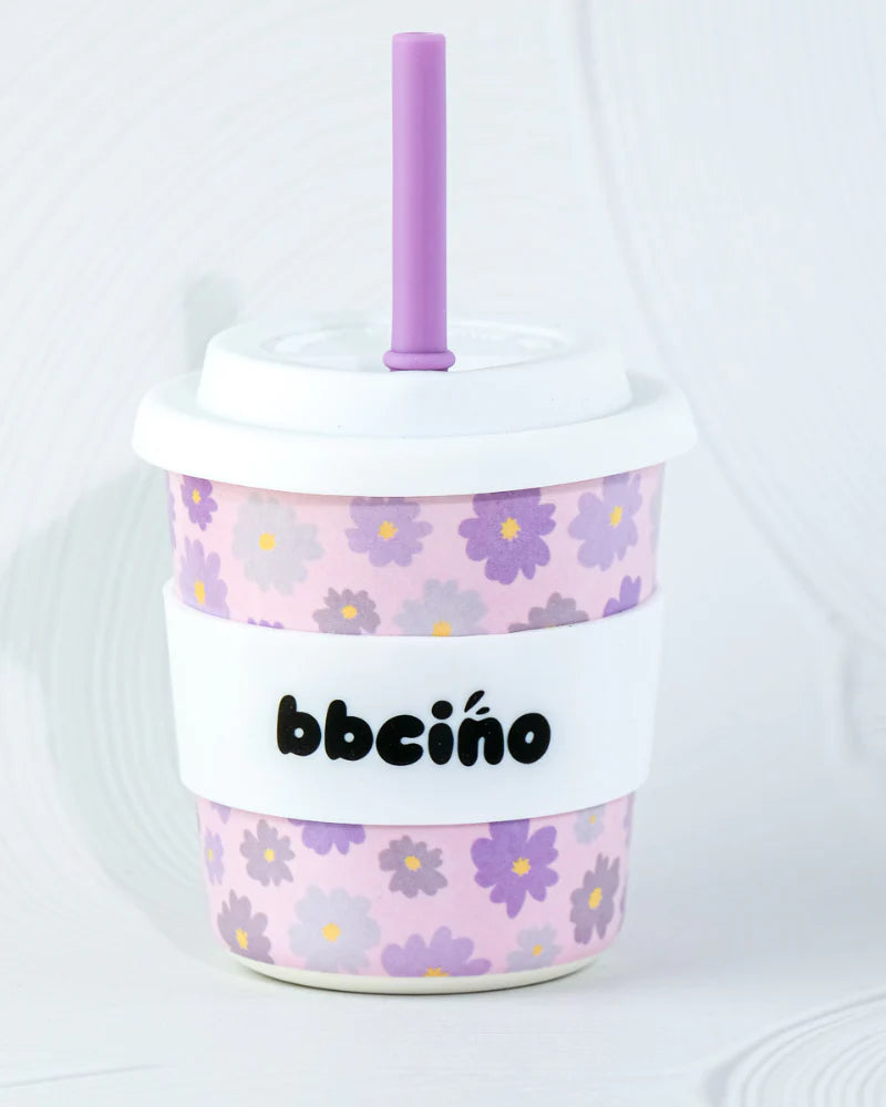 The BBcino ~ Reusable BambinoCino Cup 240ml Blossom by BBCINO is an eco-friendly toy cup made from sustainably farmed bamboo. It showcases a purple floral pattern with a purple straw, featuring a white lid and band with the text "bbciño," set against a plain, light-colored background.