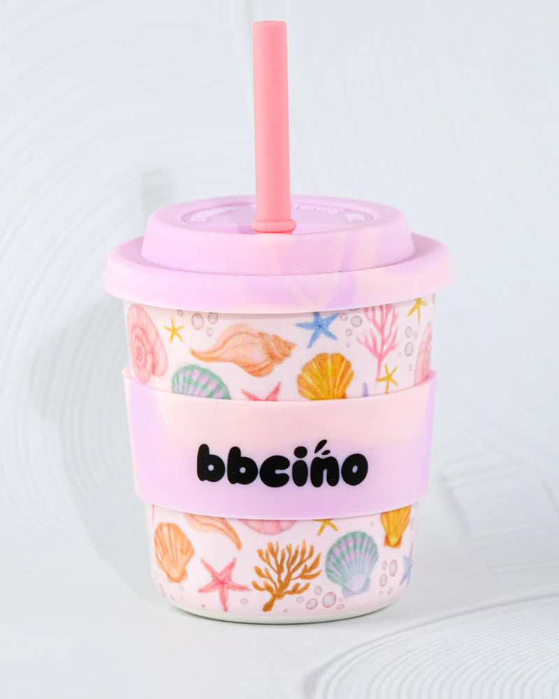 The BBcino Reusable BambinoCino Cup 240ml Shell, Yeah features seashell and starfish designs with a pink lid and straw. Made from eco-friendly materials, it includes a pink "bbciño" sleeve against a soft gradient background.