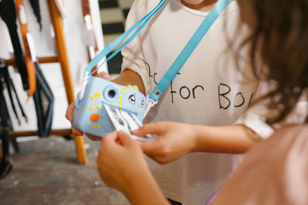 Two partially visible children are using the blue Oh My Frankie - Oh My Snap Instant Print Camera from OH MY FRANKIE, decorated with stickers. One child holds an emerging instant photo in an indoor setting filled with various items that spark creativity.