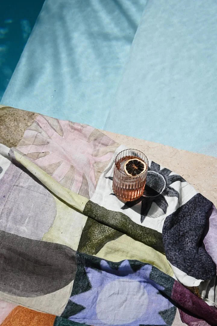 The MILKY DESIGNS Sun Checkers Wrap, featuring exclusive Annie Everingham prints, is placed poolside in sunlight with a pink glass and dried citrus slice resting on it.