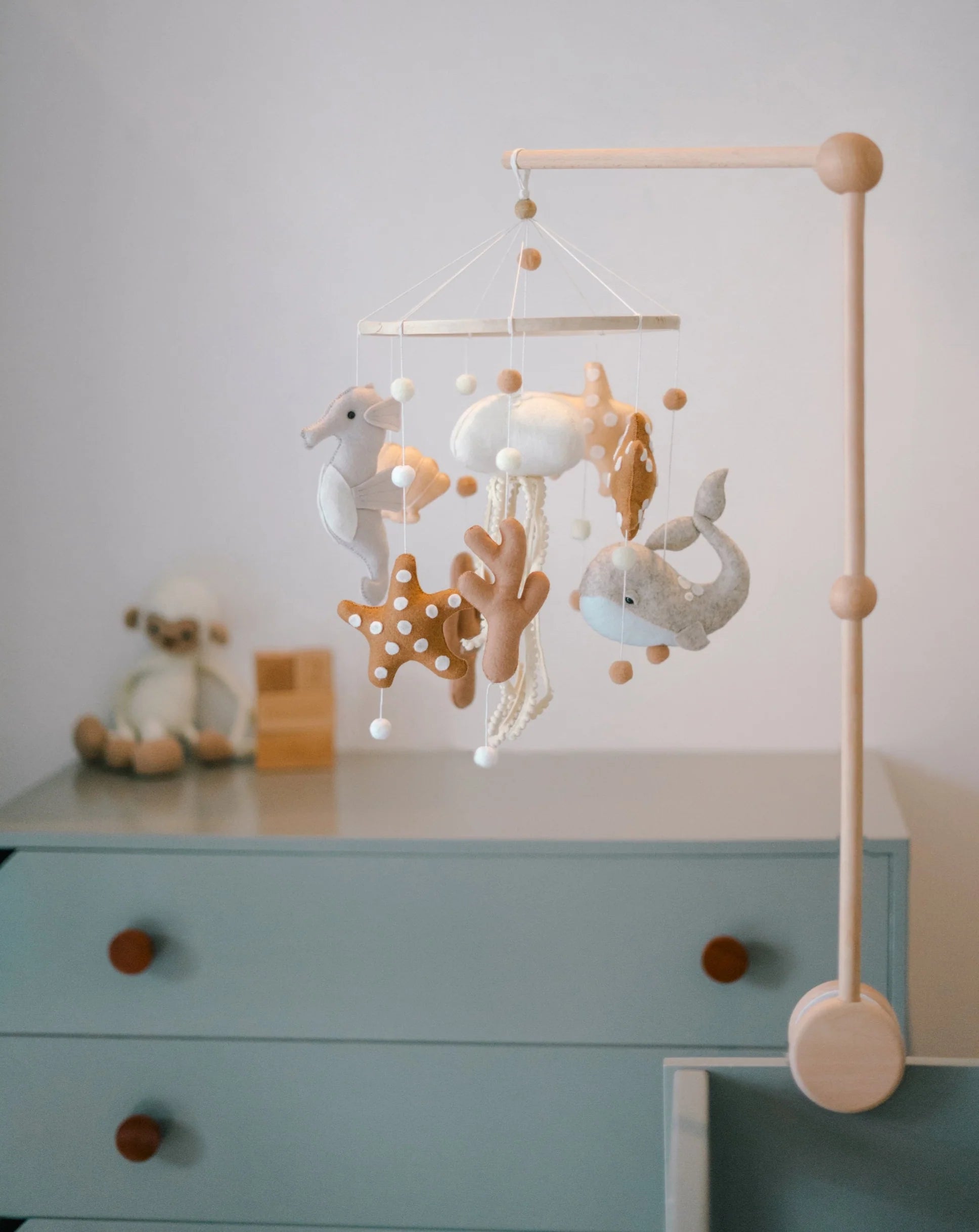 The ROOMI & ME Baby Mobile Underwater Dreaming is a handmade nursery decor featuring plush sea creatures—seahorse, starfish, whale, and jellyfish—designed for visual stimulation; shown above a blue dresser with a stuffed animal nearby.