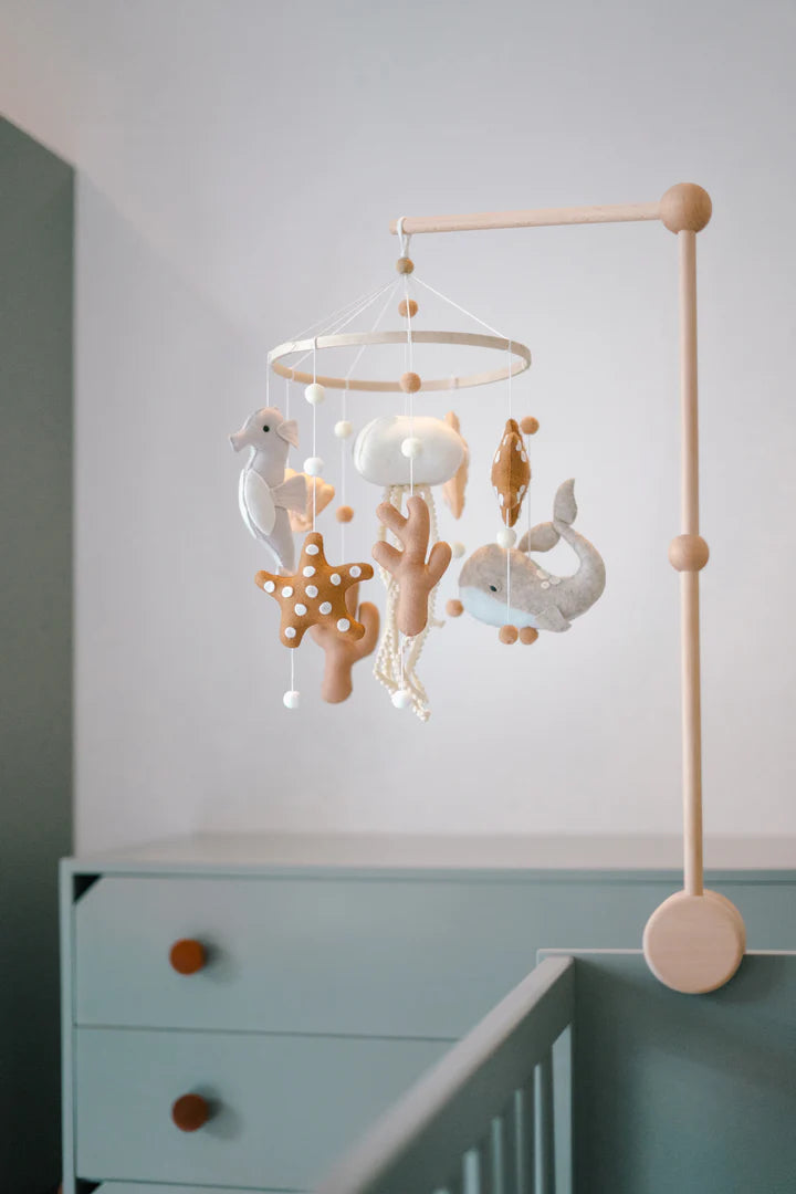 The ROOMI & ME "Roomi & Me - Baby Mobile Underwater Dreaming" features plush pastel sea creatures—a seahorse, starfish, fish, and octopus—hanging above a crib for gentle visual stimulation. A green dresser with round wooden knobs sits behind.