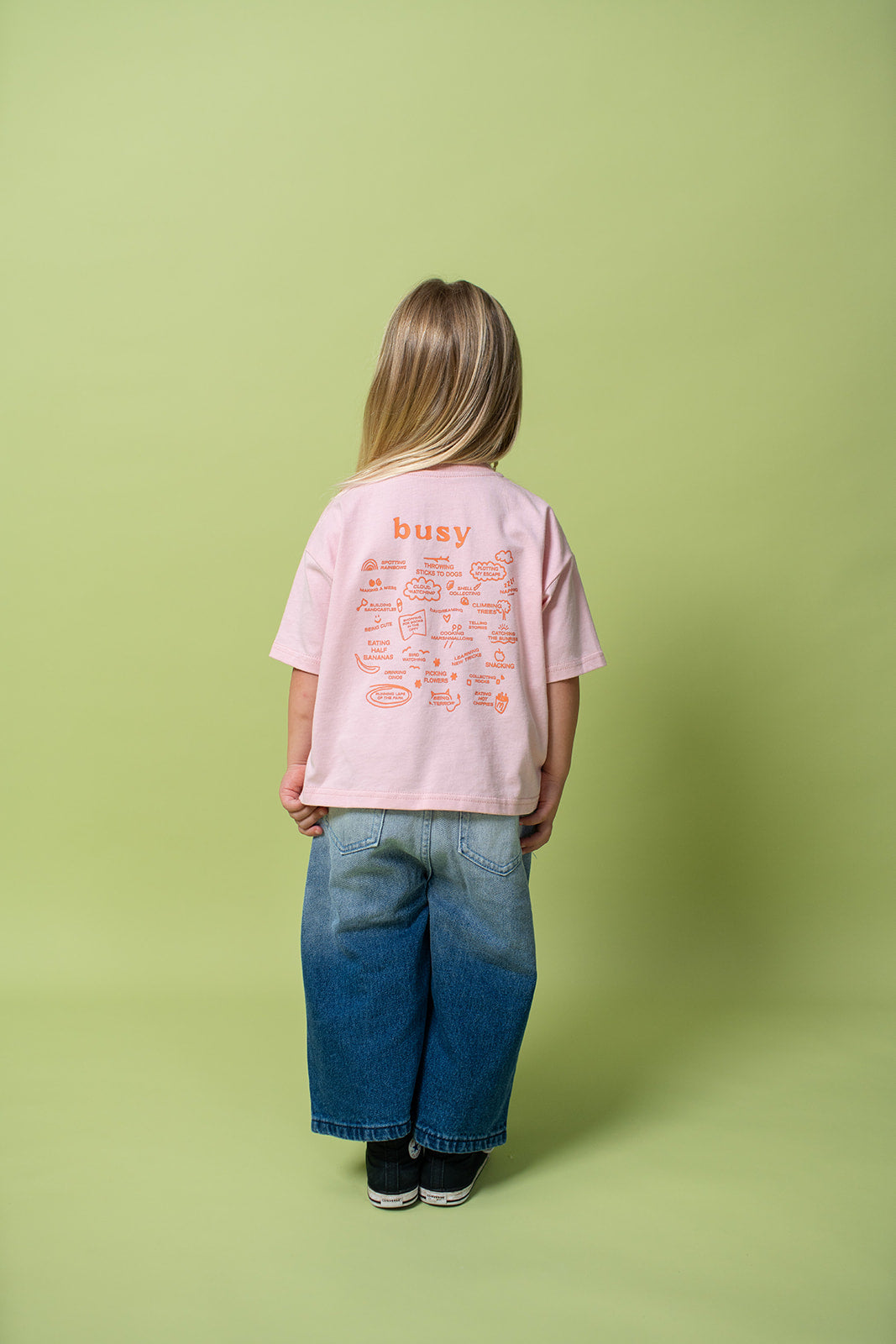 A young kid wearing the Lenn busy tee sunset shirt in pink from the back.