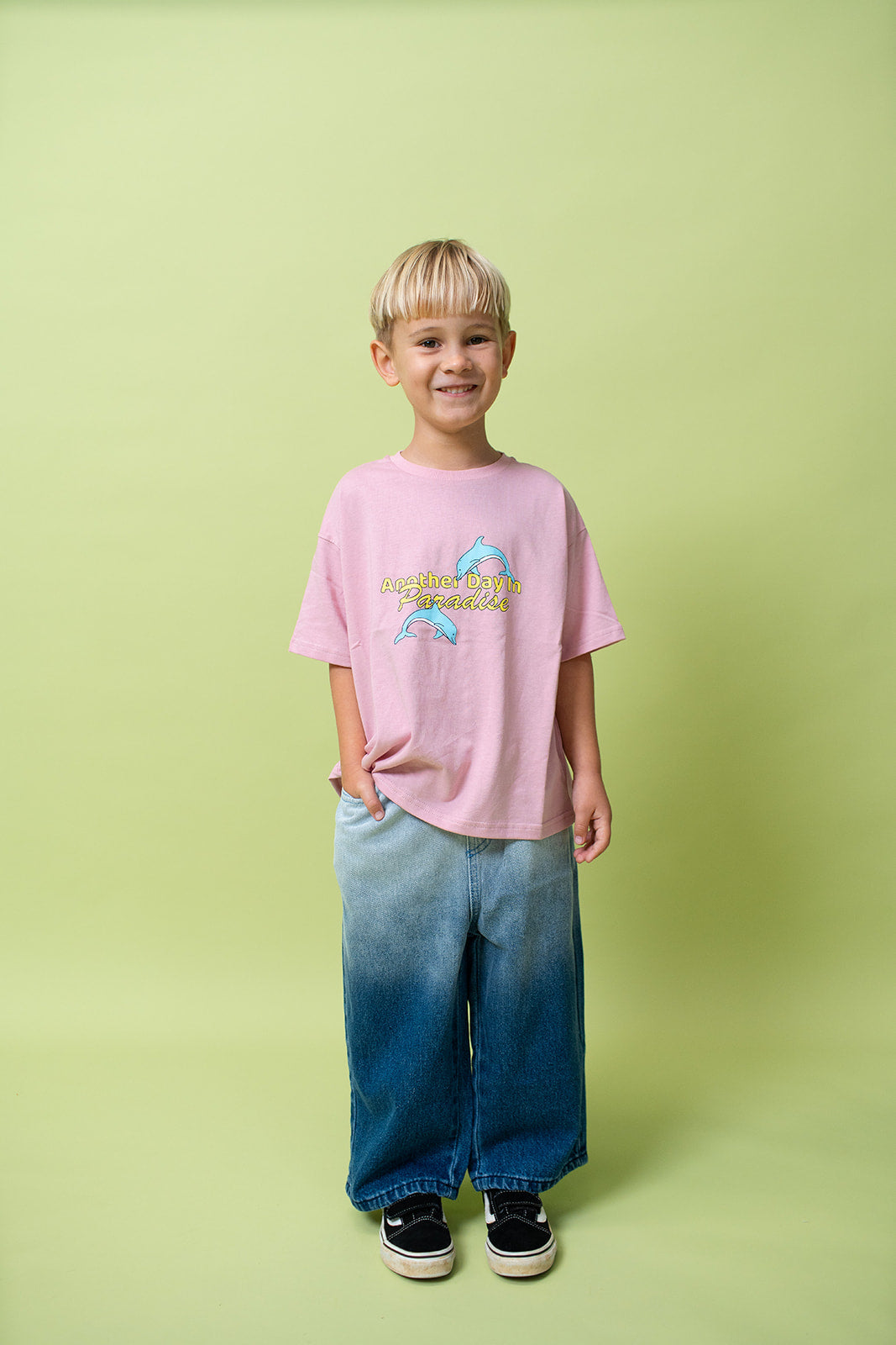 A young kid wearing the Lenn paradise tee in musk wearing jeans and black shoes with his hand in his pocket.