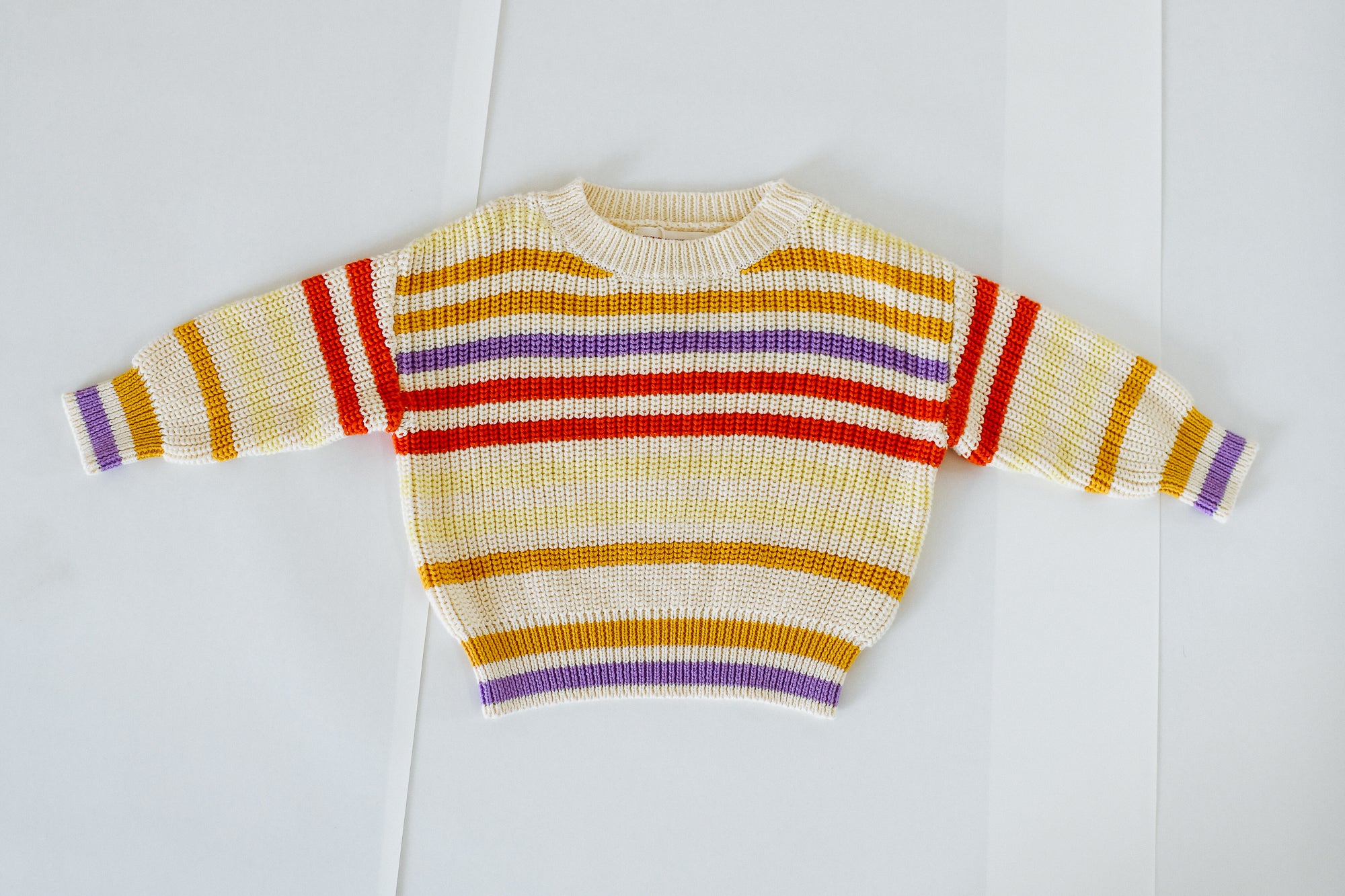 The Sunday Siblings - Art Club Knit by SUNDAY SIBLINGS is a 100% cotton sweater featuring a striped knit in yellow, orange, red, and purple on white, shown laid flat on a white surface.