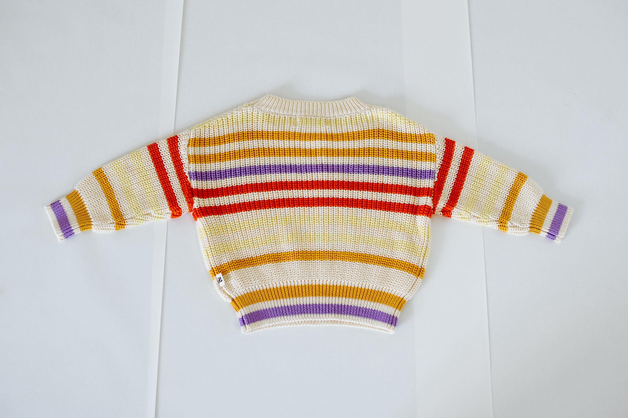 The SUNDAY SIBLINGS Sunday Siblings - Art Club Knit is a 100% cotton sweater with orange, yellow, red, and purple stripes on white, shown laid flat with sleeves outstretched on a white surface.