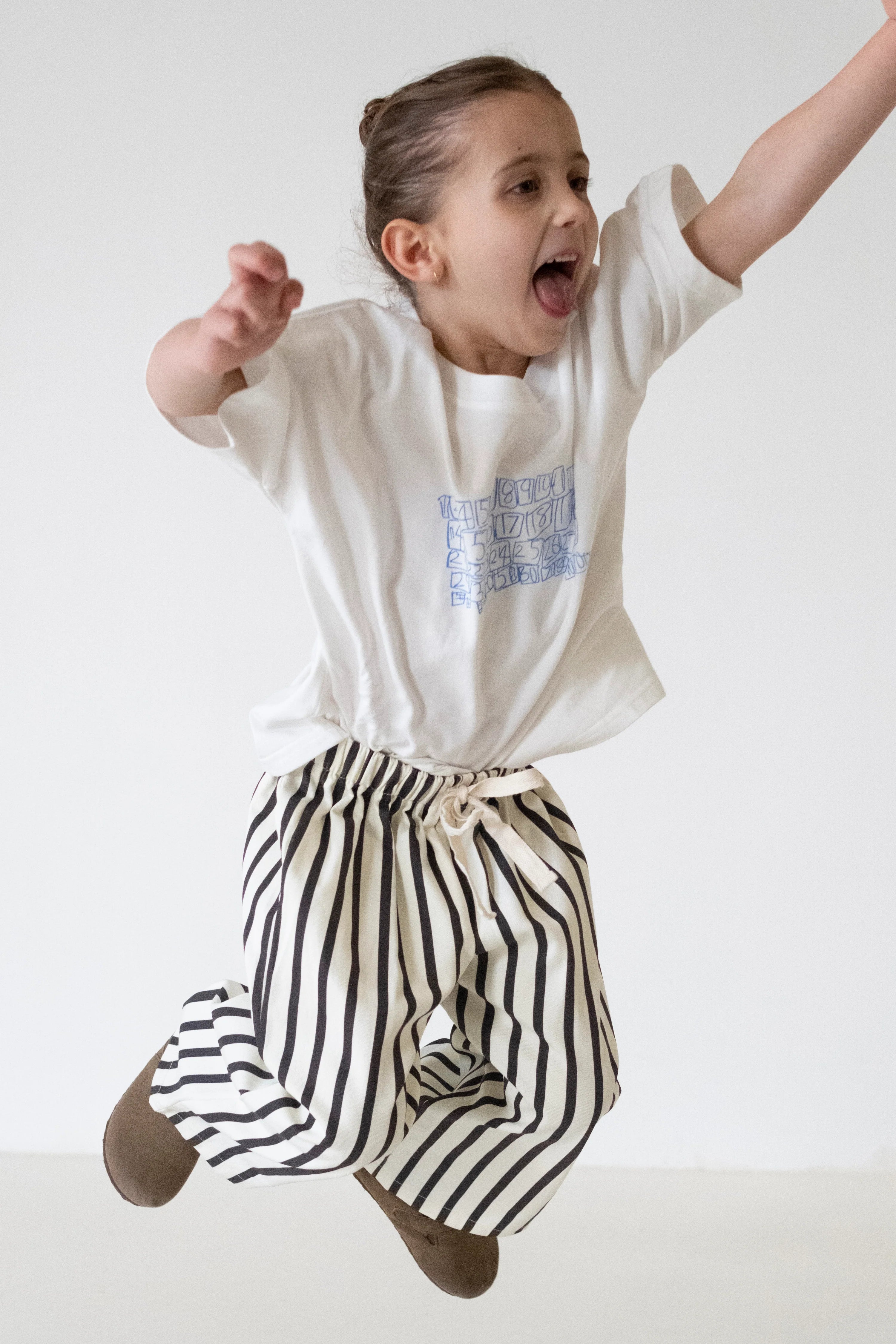 A young child with brown hair in a bun, wearing a white t-shirt and TINY TROVE Billie Stripe Cotton Pants Cream/Cocoa Stripe, jumps joyfully with arms raised against a plain light background.