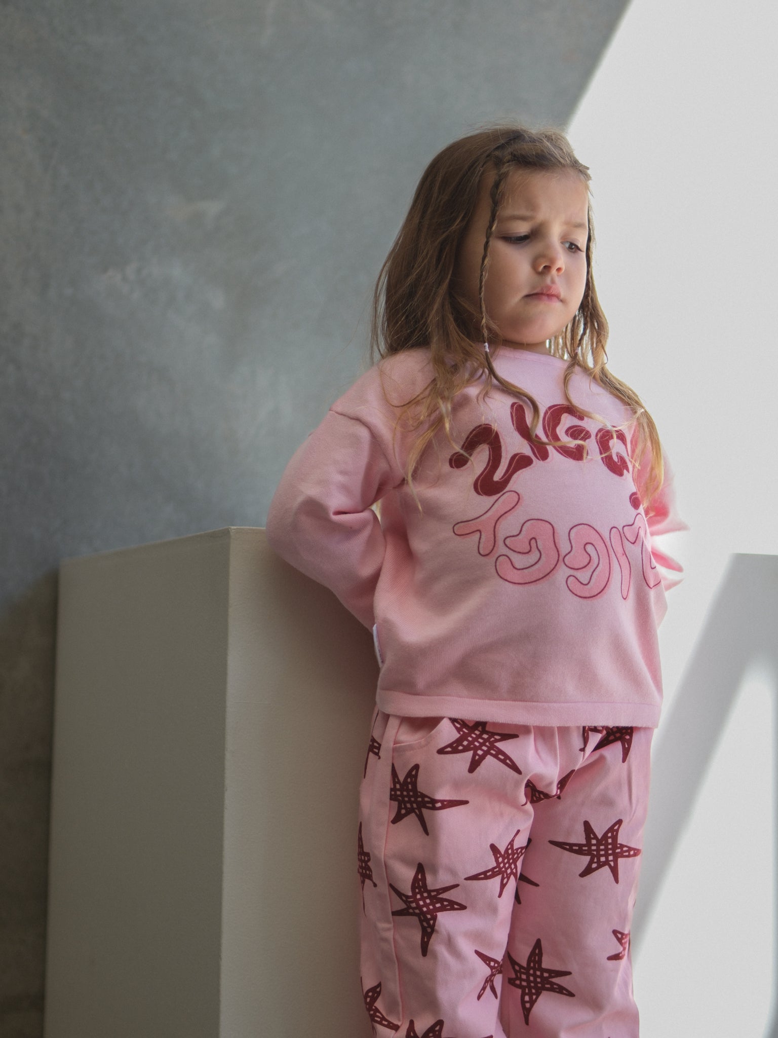 A young girl with long hair stands indoors, looking thoughtful in the ZIGGY LOU Long Sleeve Tee Thea and matching pants with red starfish patterns, her hands behind her back.