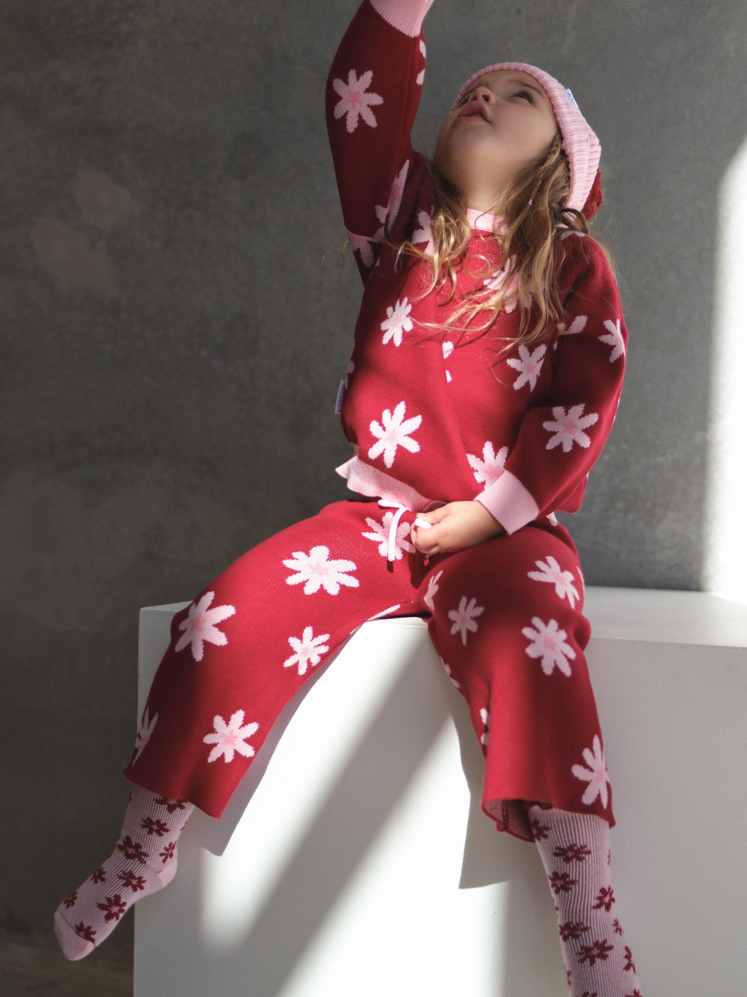 A young child in ZIGGY LOU’s Ziggy Lou - Cropped Pants Lola, a red floral outfit with matching socks and a pink hat, sits on a white block, reaching upward with their mouth slightly open.