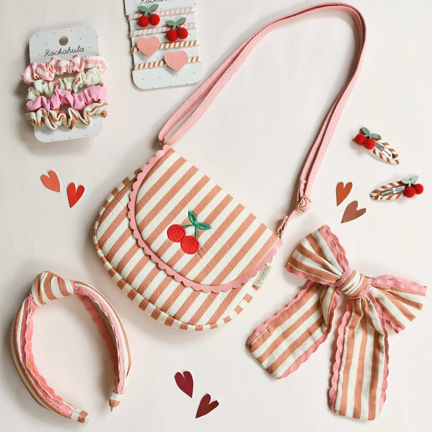 A flat lay of ROCKAHULA’s Rockahula - Stripy Cherry Ponies and other pink and beige striped accessories, including a purse, bow, headband, scrunchies, cherry hair elastics, and heart cutouts on a white background.