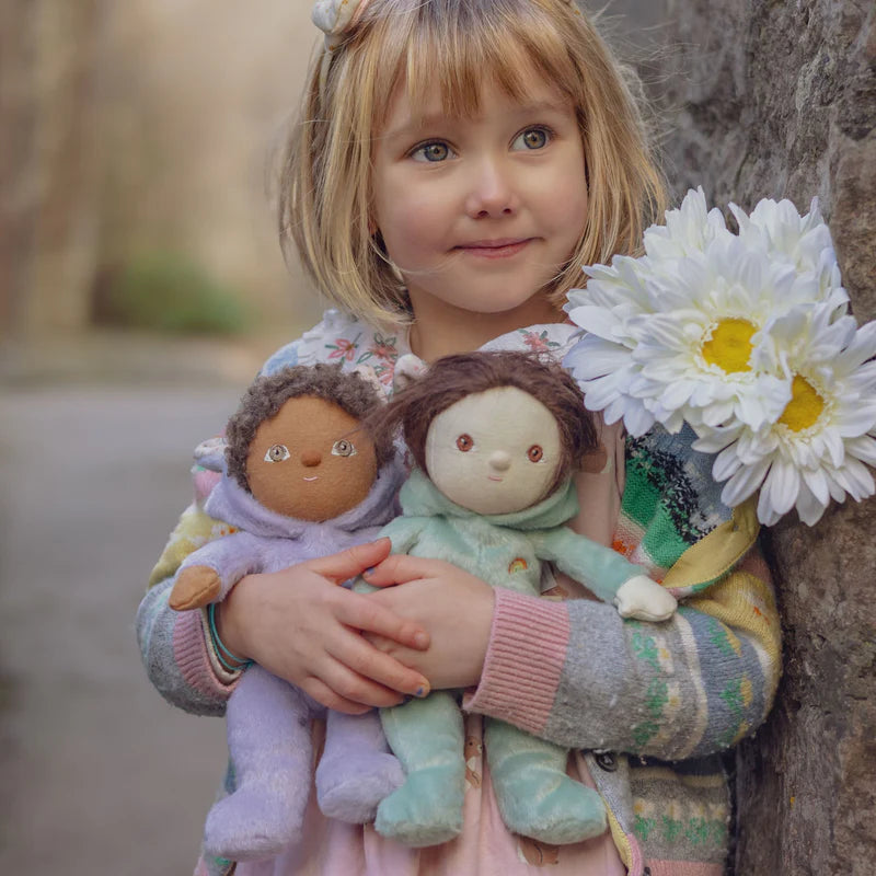 A young girl with short blonde hair holds two Olli Ella soft dolls from the Dinky Series. One doll has brown skin and curly hair, while the other has light skin and straight hair. She wears a colorful sweater and holds white flowers, standing outdoors against a blurred background.