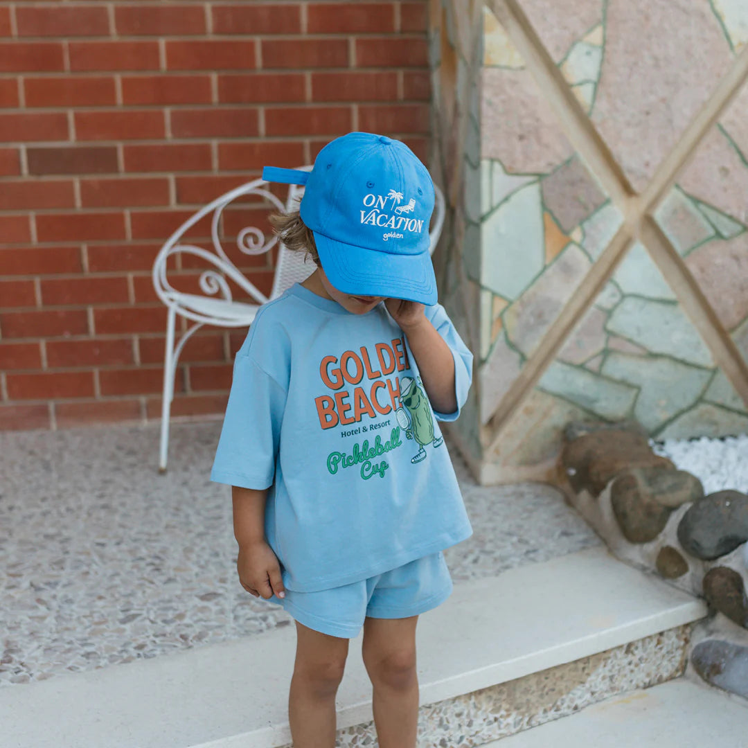 A child in a bright blue cap and cotton jersey outfit stands outdoors, covering their face. The T-shirt reads "Golden Beach" with a small illustration. They are by a white metal chair and stone-tiled wall, wearing GOLDEN CHILDREN's Retro Boy Shorts Palm Blue with an elasticated waist.