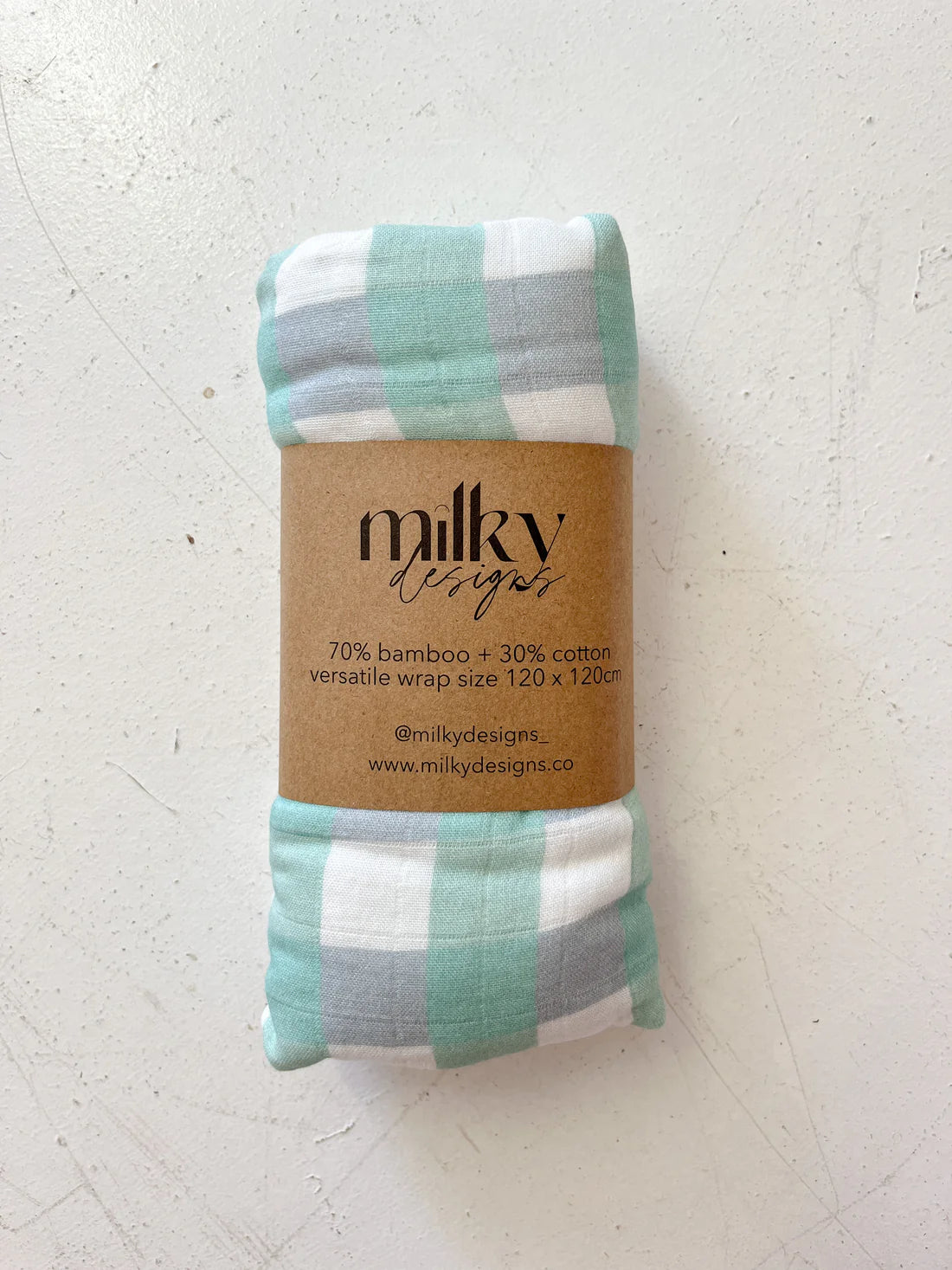 The Milky Designs ~ Seamist Gingham Wrap, by MILKY DESIGNS, is a 120 x 120 cm rolled-up fabric wrap with a green, white, and gray plaid pattern. It's secured with a brown paper band labeled "milky designs," offering buttery softness and composed of 70% bamboo and 30% cotton.