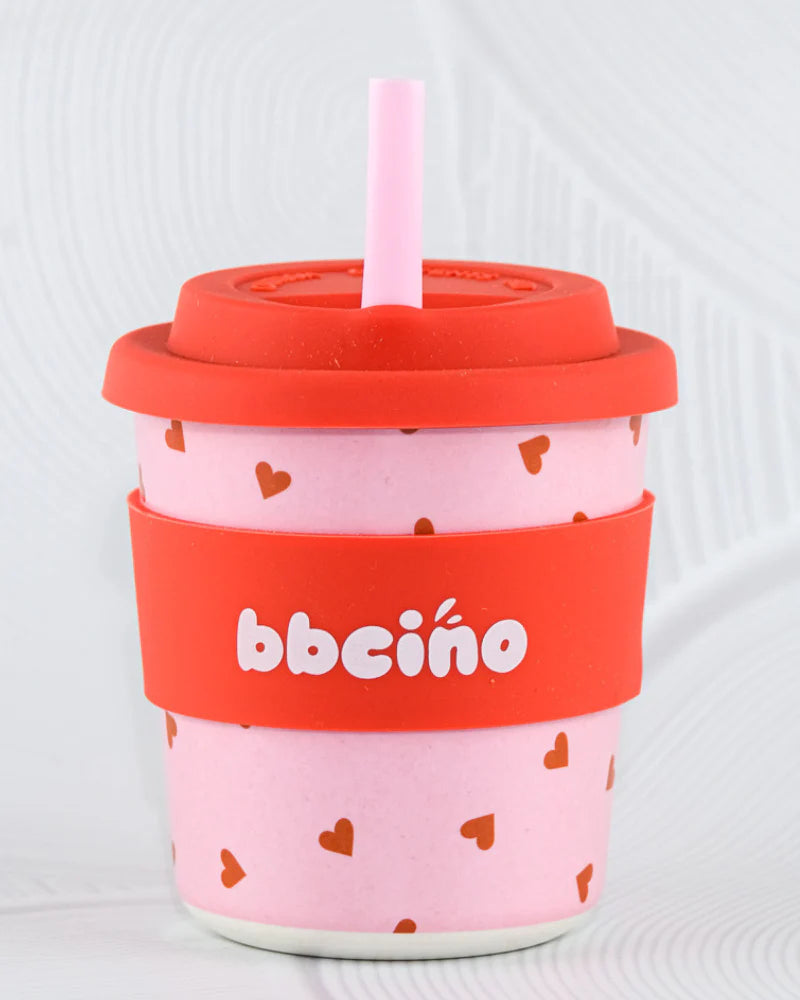 The BBcino Reusable BambinoCino Cup 240ml Sweetheart by BBCINO features pink bamboo, red heart patterns, a red "bbcino" silicone sleeve, matching lid, and light pink straw. Eco-friendly with a light textured background.