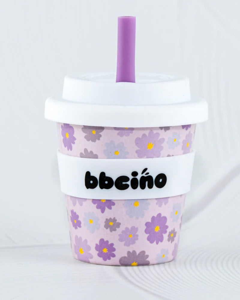 The BBcino Reusable Babycino Cup 120ml Blossom by BBCINO features a white lid, purple straw, and purple-yellow floral design. Made from eco-friendly materials with a "bbceíno" band, it's perfect for reusable cup lovers.