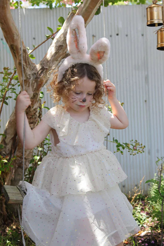 A child in a cream dress with pastel polka dots stands on a swing, wearing the OLLI ELLA Fluffle Bunny Ear Headband in pink. Whiskers drawn on their cheeks add to the playful scene set against a tree and corrugated metal fence backdrop.