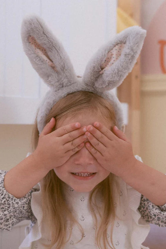 A young child with long blonde hair covers their eyes with both hands while wearing a white patterned top and the Olli Ella ~ Fluffle Bunny Ear Headband Smoke by OLLI ELLA, complete with fluffy gray posable ears, sitting indoors.