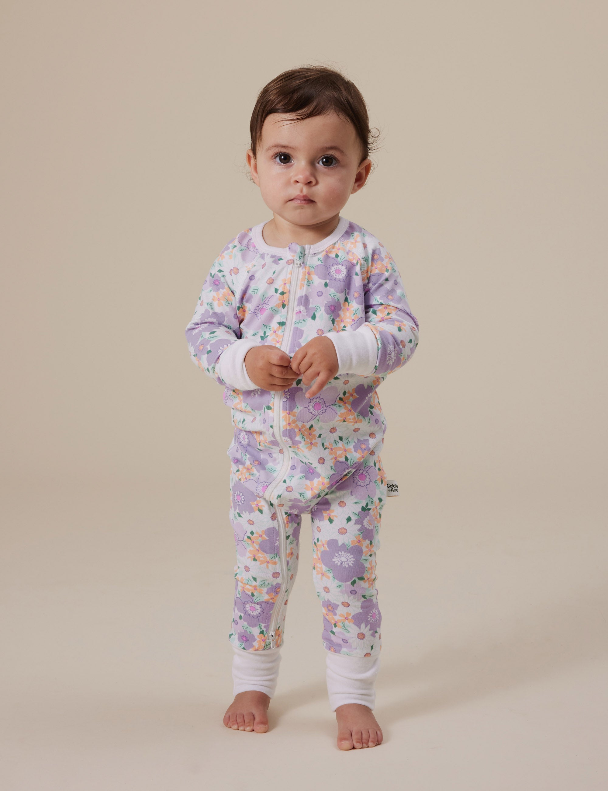 A toddler poses on a plain background, dressed in the Goldie + Ace ~ Flora Zipsuit from GOLDIE + ACE. The onesie features a pastel floral print, ribbed cuffs, and a two-way zip. The child has a neutral expression and bare feet.