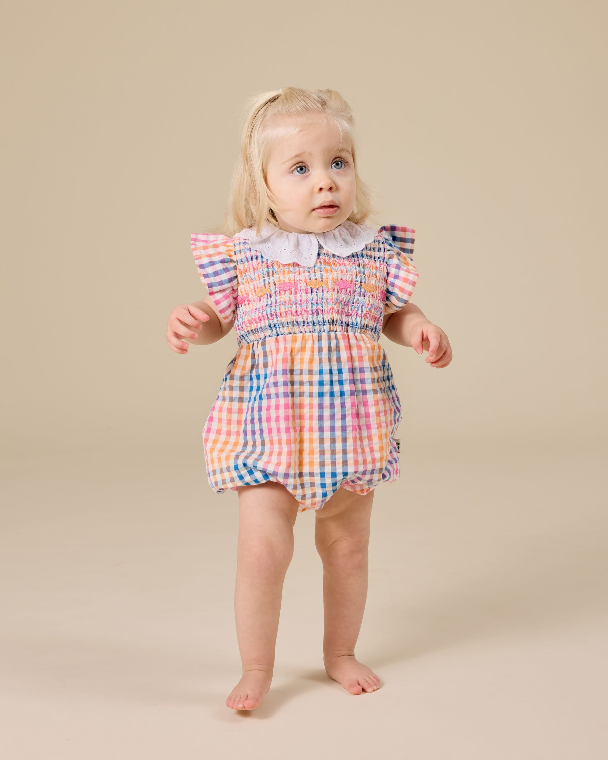 A young blonde child stands barefoot on a beige studio floor, wearing the GOLDIE + ACE Rainbow Gingham Lani Smocked Romper with an elegant broderie anglaise collar and ruffled sleeves, gazing upward in surprise.