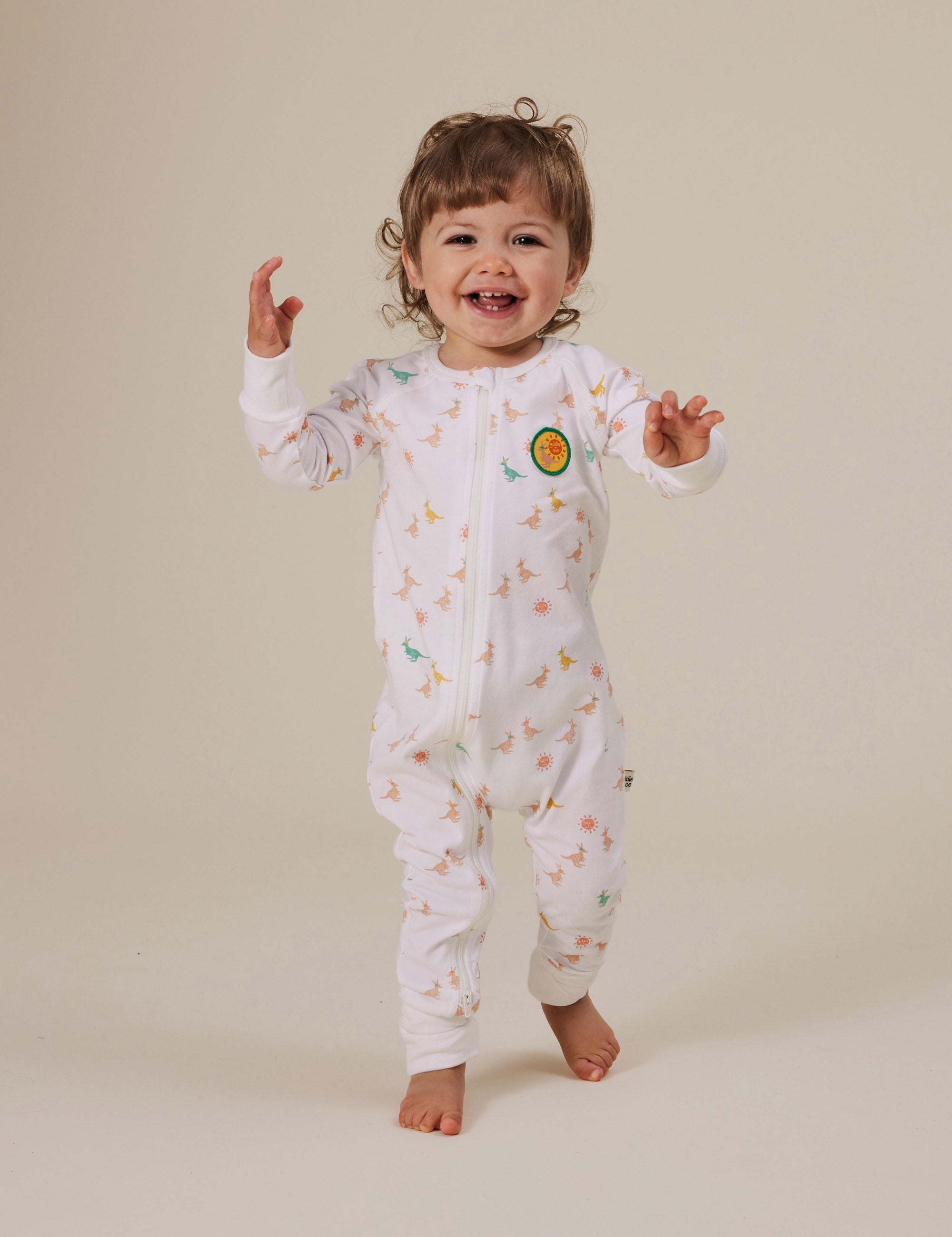 A toddler with curly hair joyfully walks forward, wearing the Goldie + Ace ~ Joey Zipsuit from GOLDIE + ACE. The white onesie features colorful dinosaur patterns and a handy two-way zip, as the child smiles broadly, balancing with arms slightly raised against a neutral background.