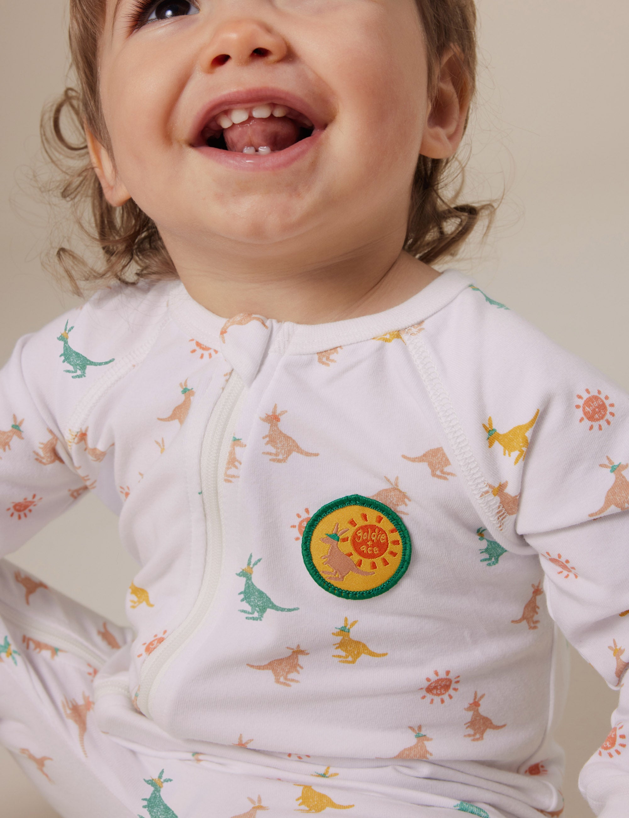 A toddler in the Goldie + Ace ~ Joey Zipsuit featuring a playful joey print of colorful dinosaurs and sun patches. The child sits contentedly, enjoying the comfort of extendable ribbed cuffs and a convenient two-way zip against a neutral background.