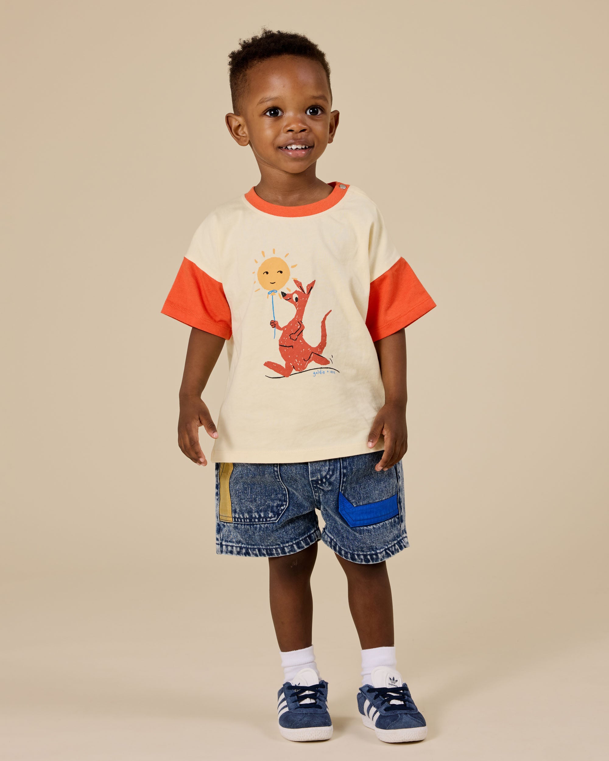 A young child smiles in a cream and orange T-shirt with a kangaroo and sun graphic, wearing GOLDIE + ACE Primary Pop Noah Denim Shorts featuring blue and yellow patches, plus blue sneakers, all set against a plain beige background.