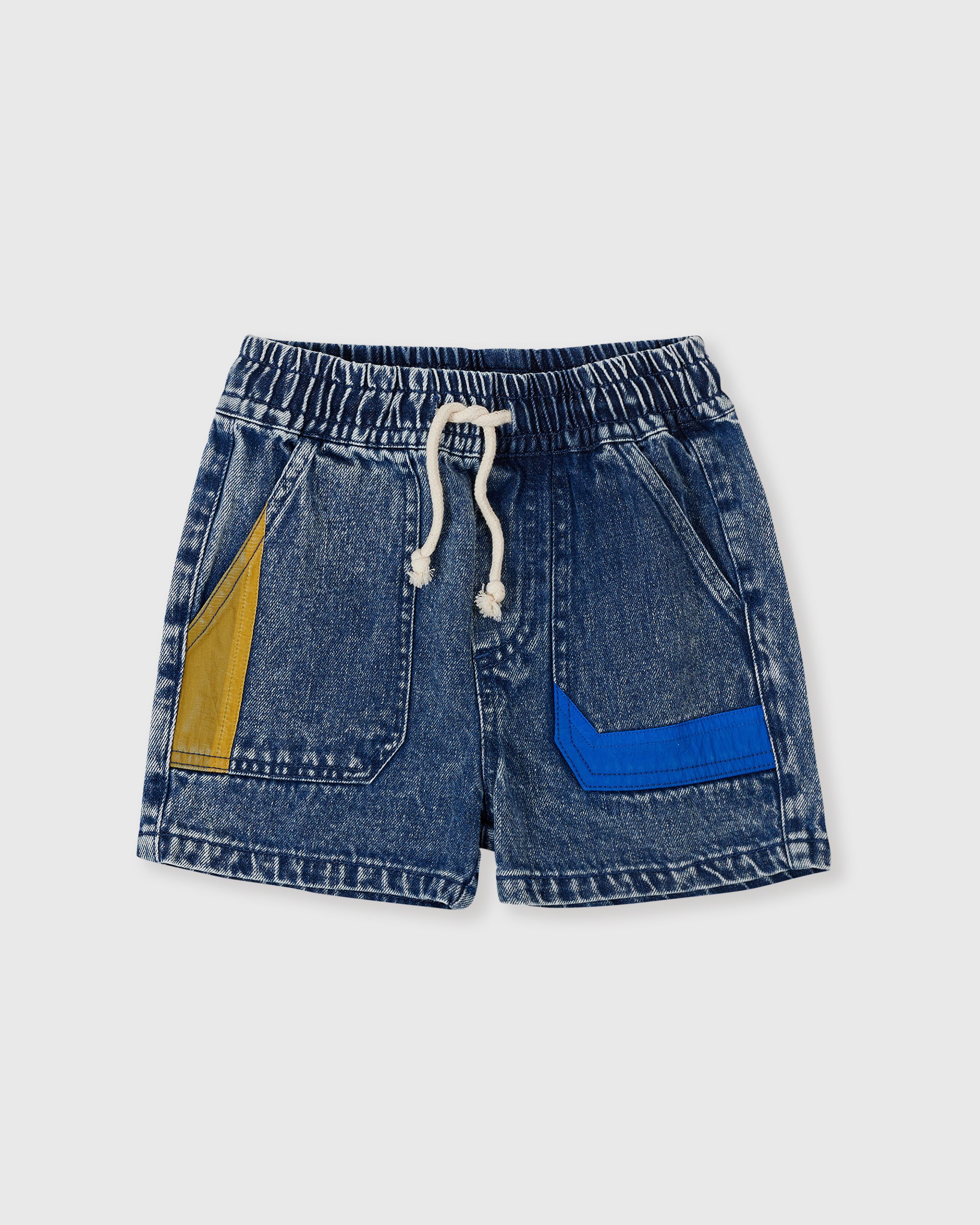 The GOLDIE + ACE Primary Pop Noah Denim Shorts feature a classic cut in blue denim with an elasticated waist, white drawstring, and two front pockets: one with a yellow patch on the left and one with a blue patch on the right. Shot laid flat on a light background.