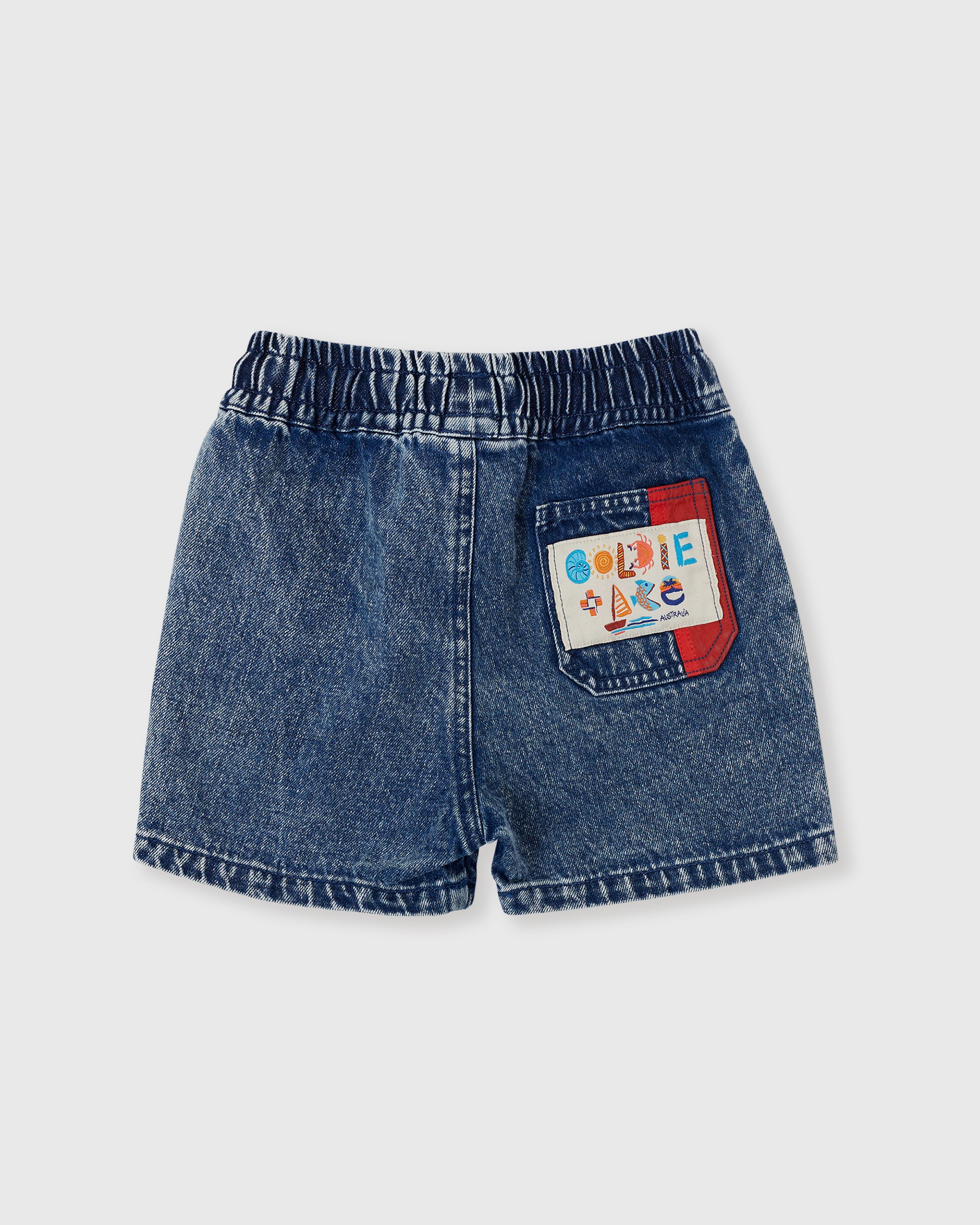 The GOLDIE + ACE Primary Pop Noah Denim Shorts feature a classic cut, elasticated waist, and a colorful patch with playful text on the back pocket, shown laid flat against a light background.