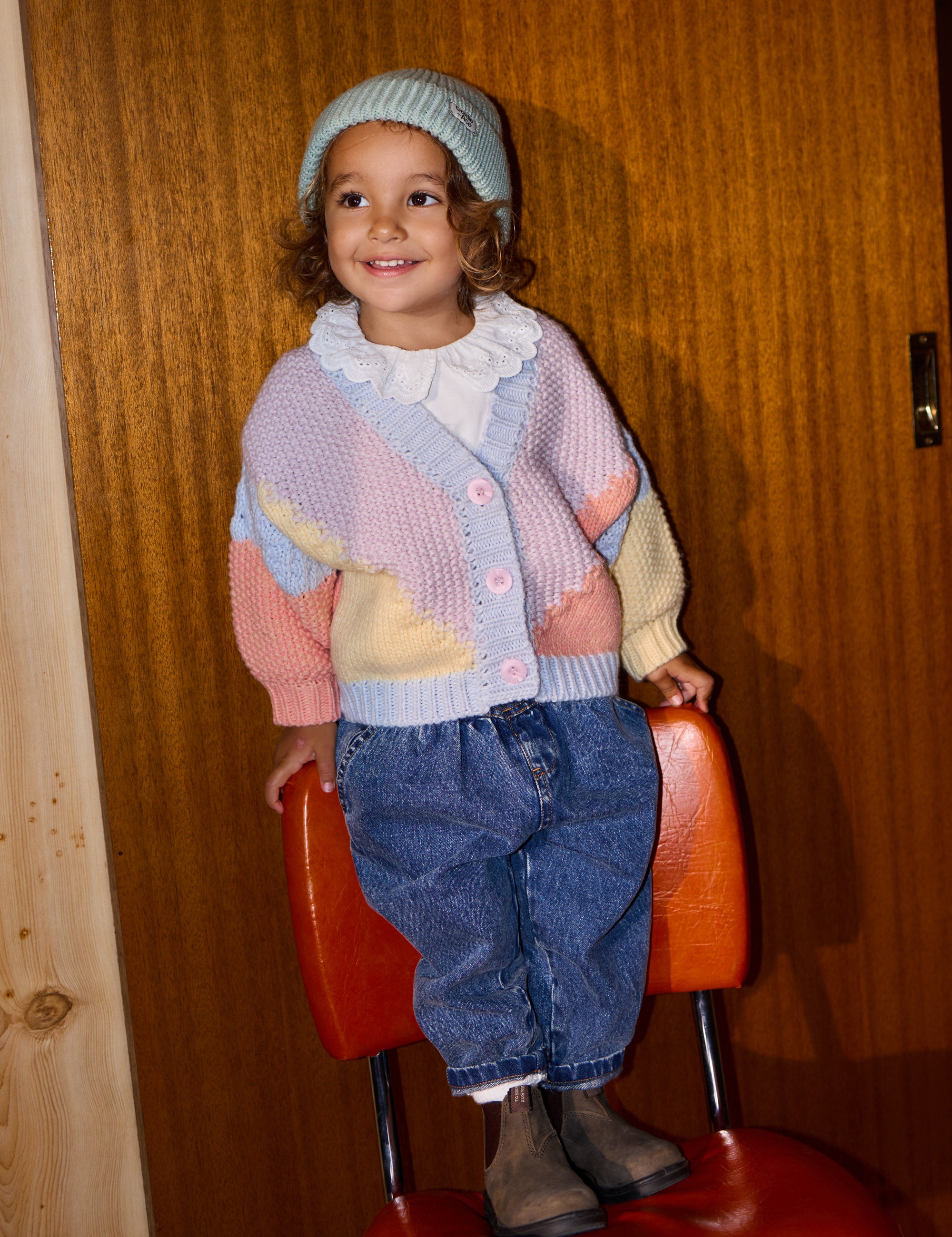 A smiling child stands on an orange chair in front of a wooden door, wearing a multicolored knitted cardigan, the Goldie + Ace ~ Wool Beanie Mint by GOLDIE + ACE, jeans, and brown boots.
