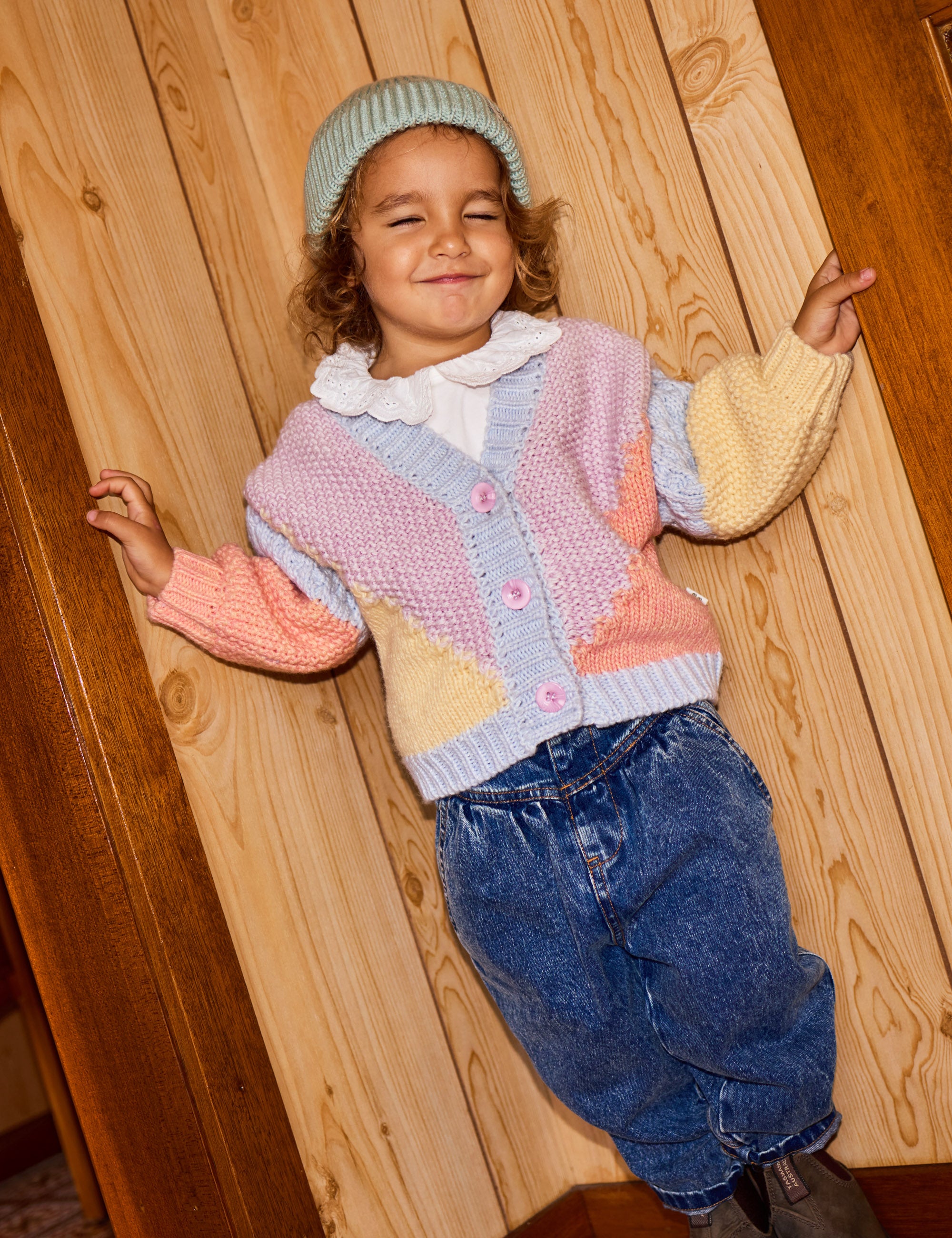 A young child stands smiling with eyes closed, wearing a pastel-colored knitted cardigan, white frilled collar shirt, denim pants, and the Goldie + Ace ~ Wool Beanie Mint. They lean against wooden paneling, appearing cheerful and relaxed.