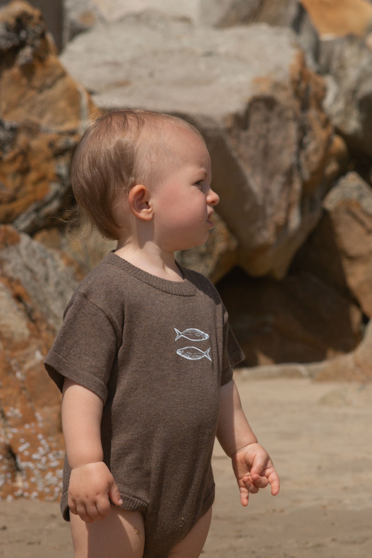 Child wearing a brown romper with fish designs standing on a rocky beach.