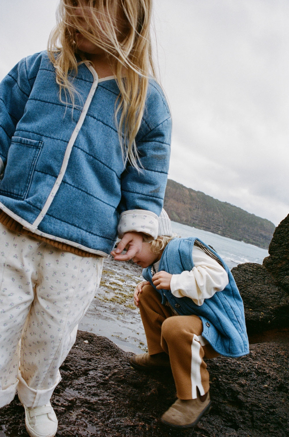 Two children in cozy layers—a blue jacket and GROWN Everyday Pants Garden, the other in a blue vest and brown pants—stand and crouch on a rocky shore, with sea and cliffs in the background beneath a cloudy sky.