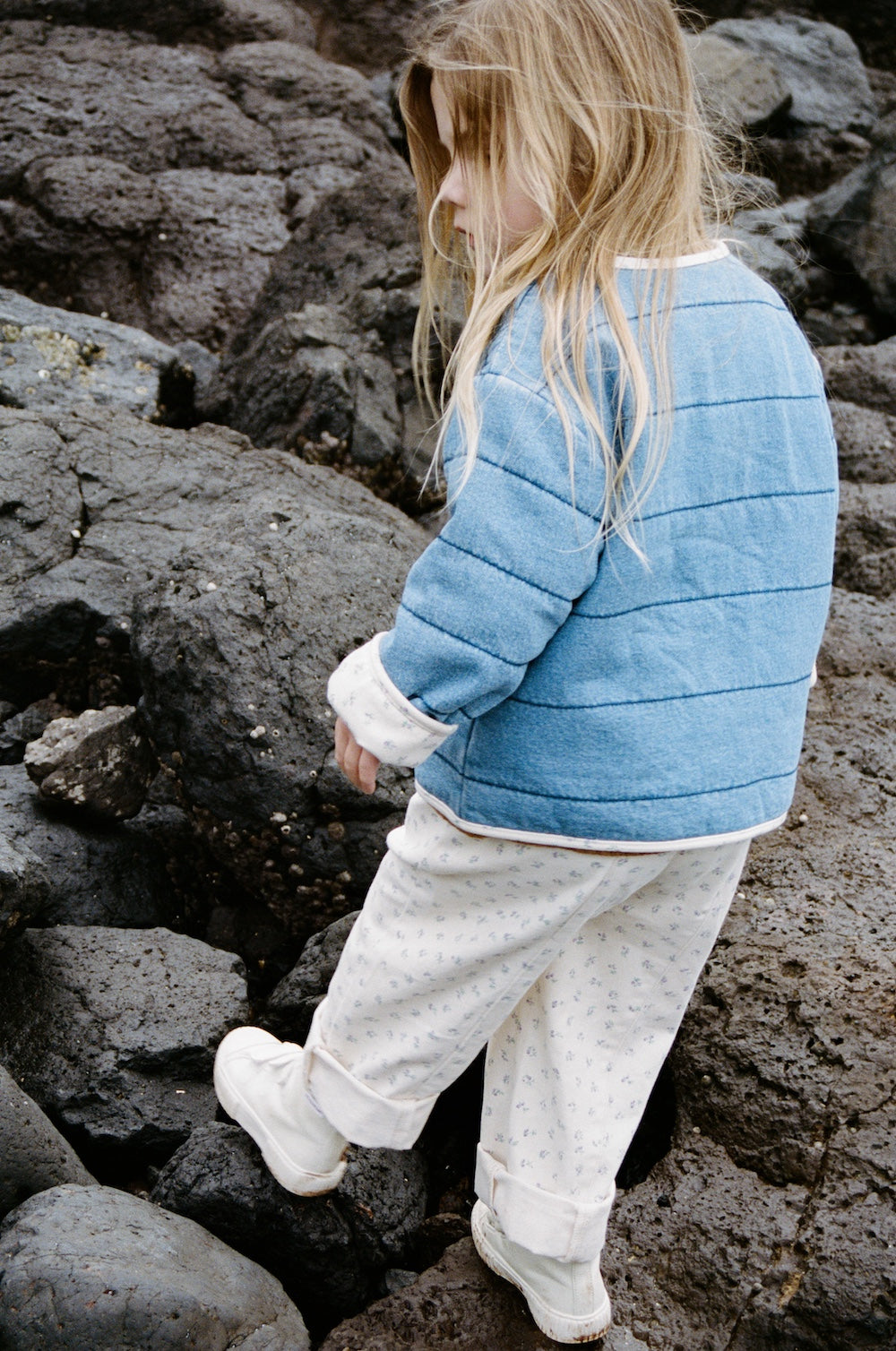 A young child with long blond hair, wearing a blue jacket and GROWN’s Everyday Pants Garden by GROWN, featuring a cotton elastic waistband, walks carefully on dark, uneven rocks outdoors.