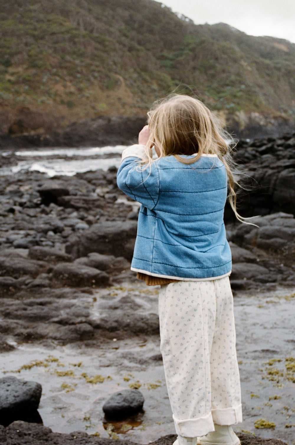 A young child with long blonde hair stands on rocky terrain near the ocean, wearing GROWN's Grown - Everyday Pants Garden and a blue jacket, gazing toward the waves and distant hills, facing away from the camera.