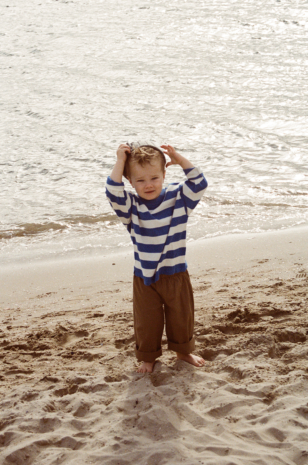 A young child in GROWN’s Organic Everyday Denim Pant Coffee stands barefoot on the sandy beach in a relaxed fit, blue and white striped long-sleeve shirt, hands on their head, looking at the camera.