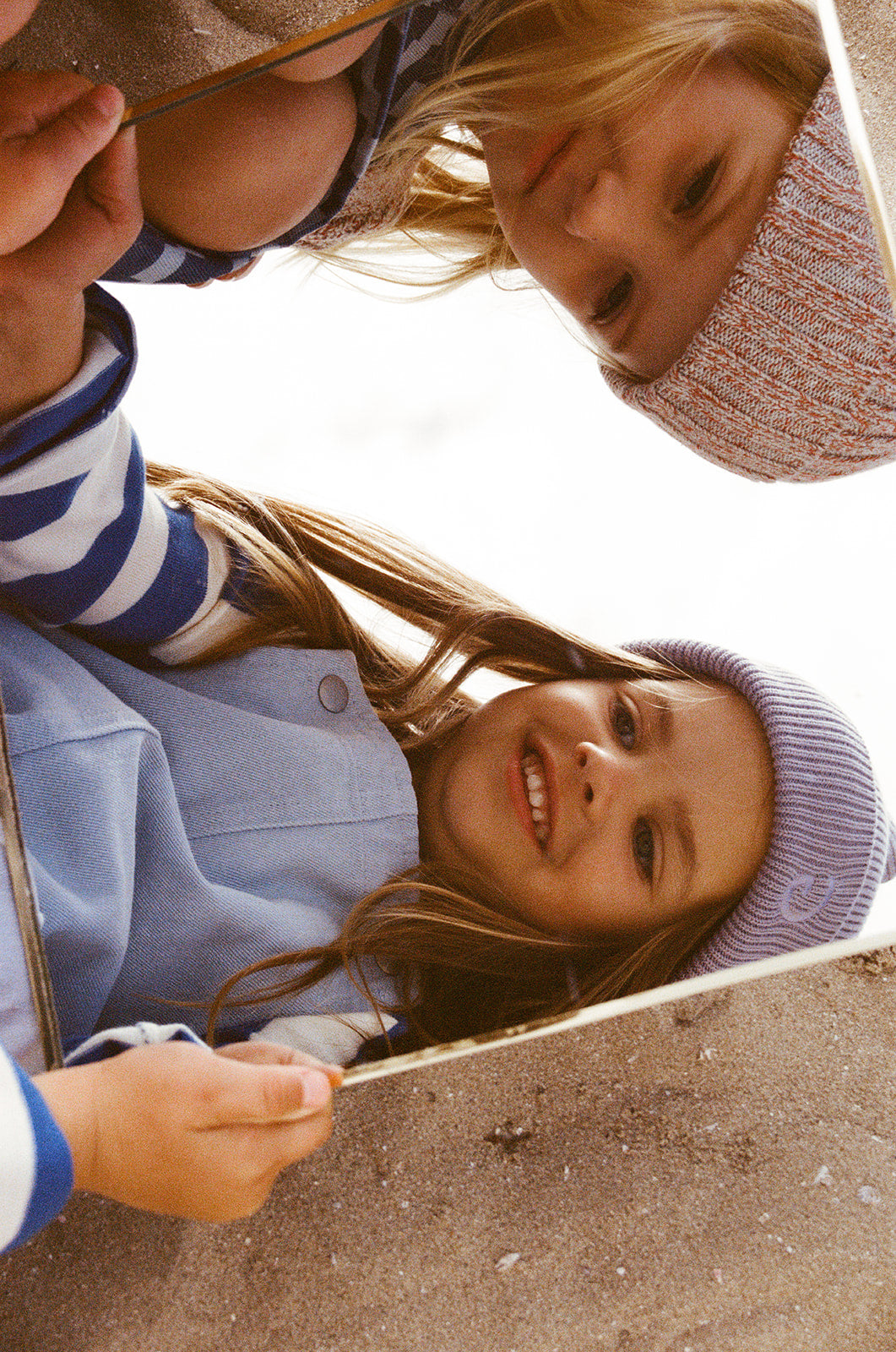 Two children in GROWN's Twisted Pixie Beanie gaze into a mirror on sandy ground, smiling. Their reflections are sharp, while their cozy outfits hint at the soft cotton and recycled polyester used by GROWN.