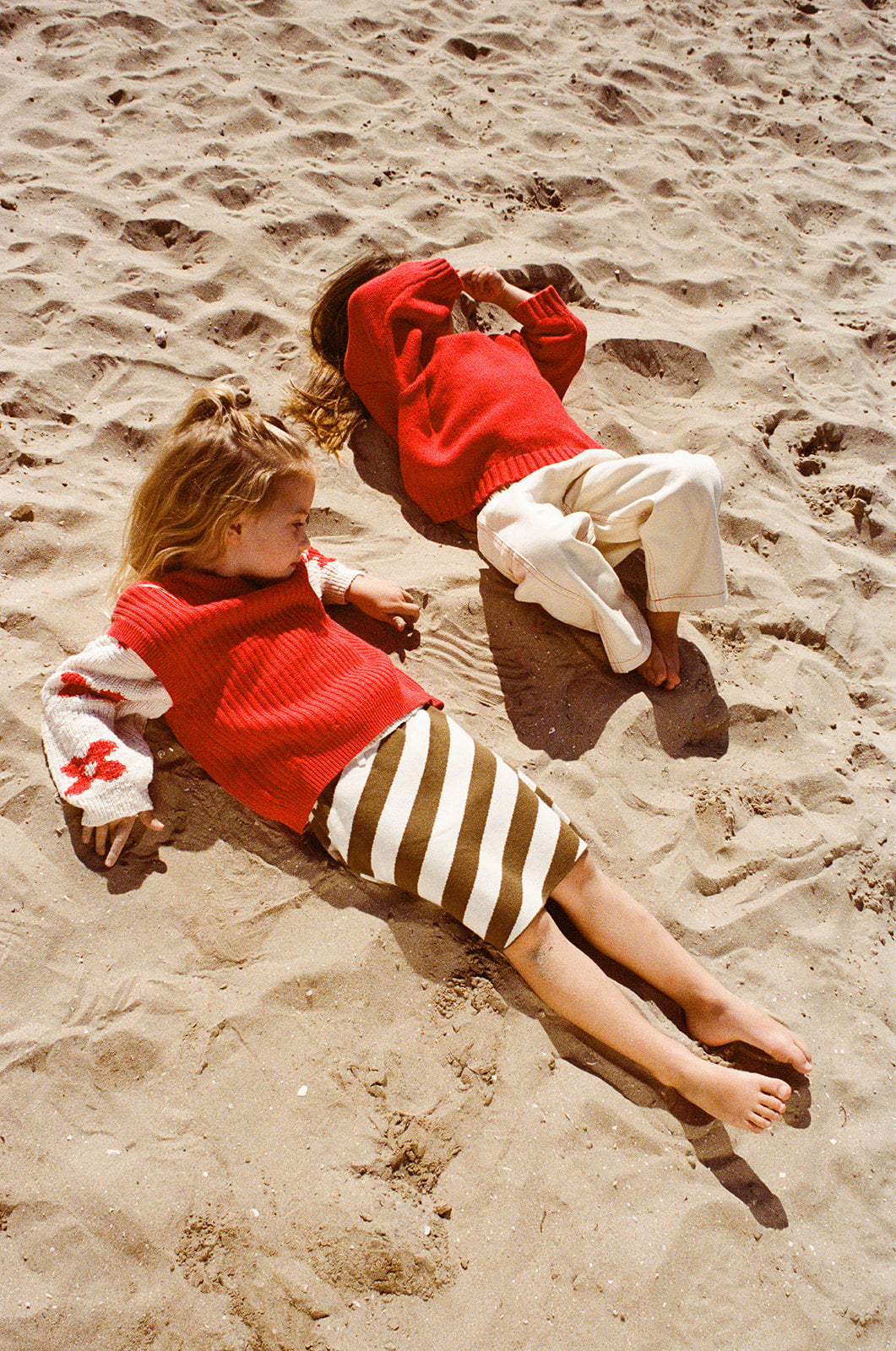 Two children lie barefoot on sand. One wears the GROWN Merino Blend Everyday Pull Over Flame sweater with cozy style; the other, in cream pants, shields their face with an arm. Both are relaxed.