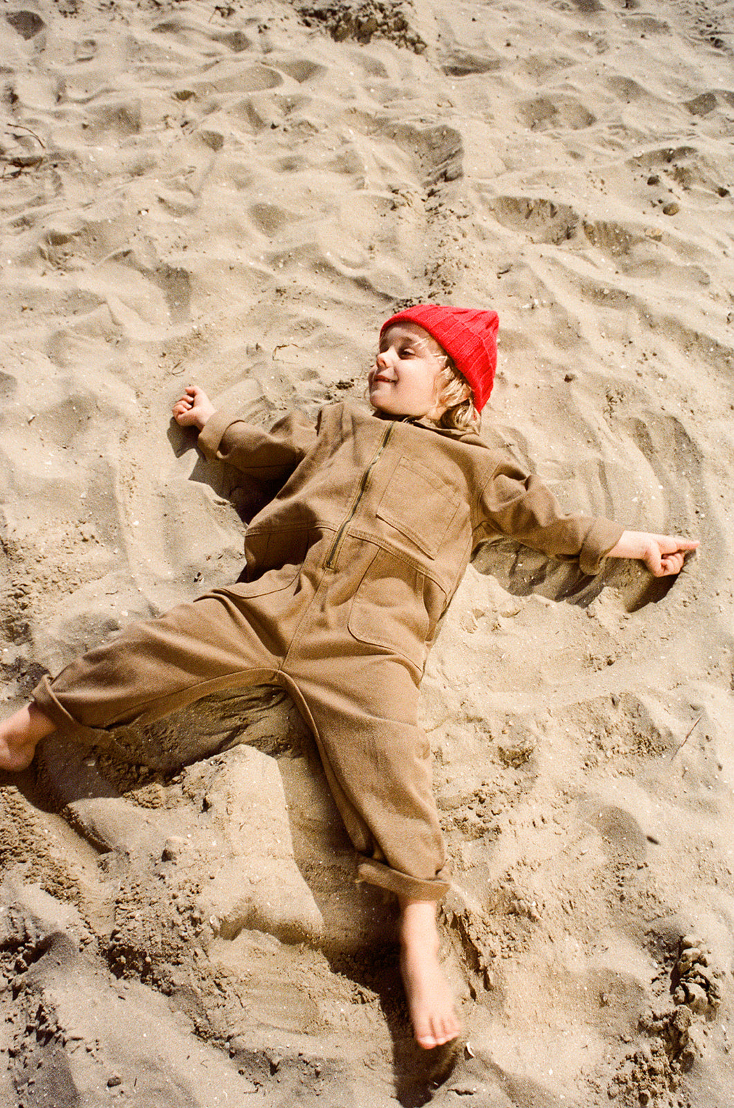 A young child wearing a brown recycled nylon jumpsuit and the GROWN Merino Blend Ribbed Beanie Flame lies on their back in the sand, smiling and spreading their arms and legs as if making a sand angel.