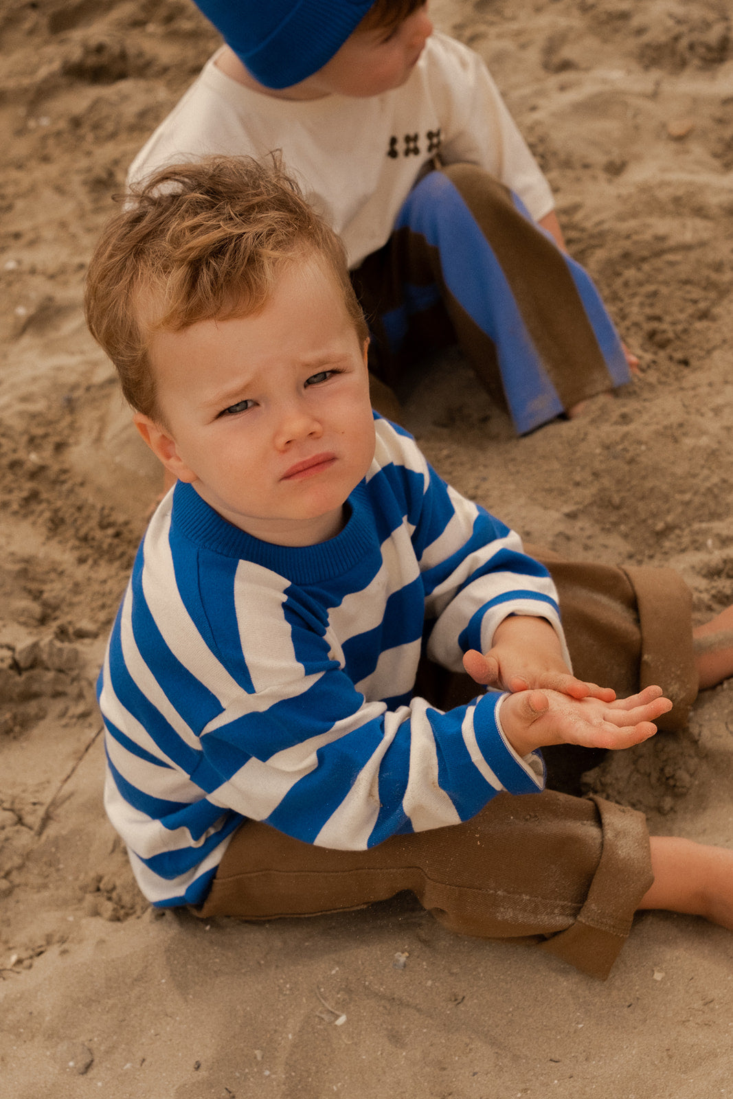 A young child wears the GROWN Stripe Long Sleeve Tee and brown pants, sitting on sandy ground with a serious look. Another child in a blue hat and white shirt is seated behind him.